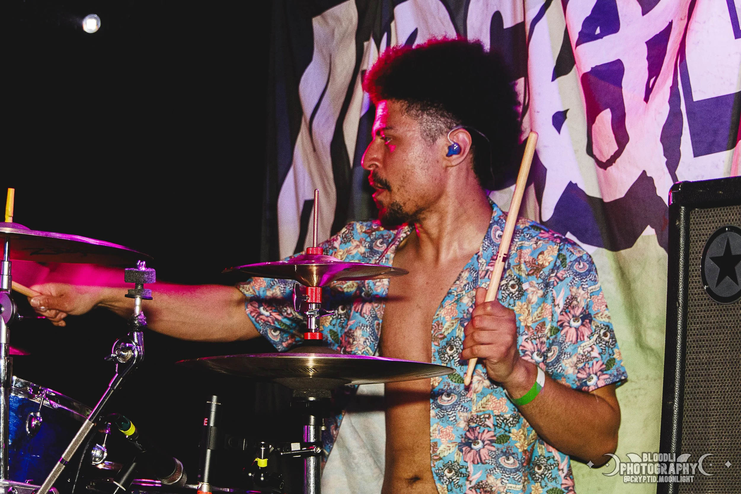 A male drummer with dark, styled hair, wearing blue in-ear monitors and a vibrant floral shirt, playing a drum set on stage with a colorful backdrop.