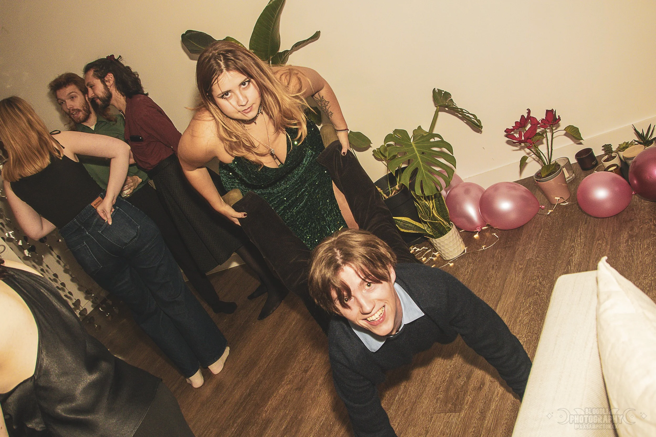 Group of friends at a party, with a young woman in a green dress sitting on a man's back, who is on all fours. The scene features pink balloons and potted plants along a plain wall.