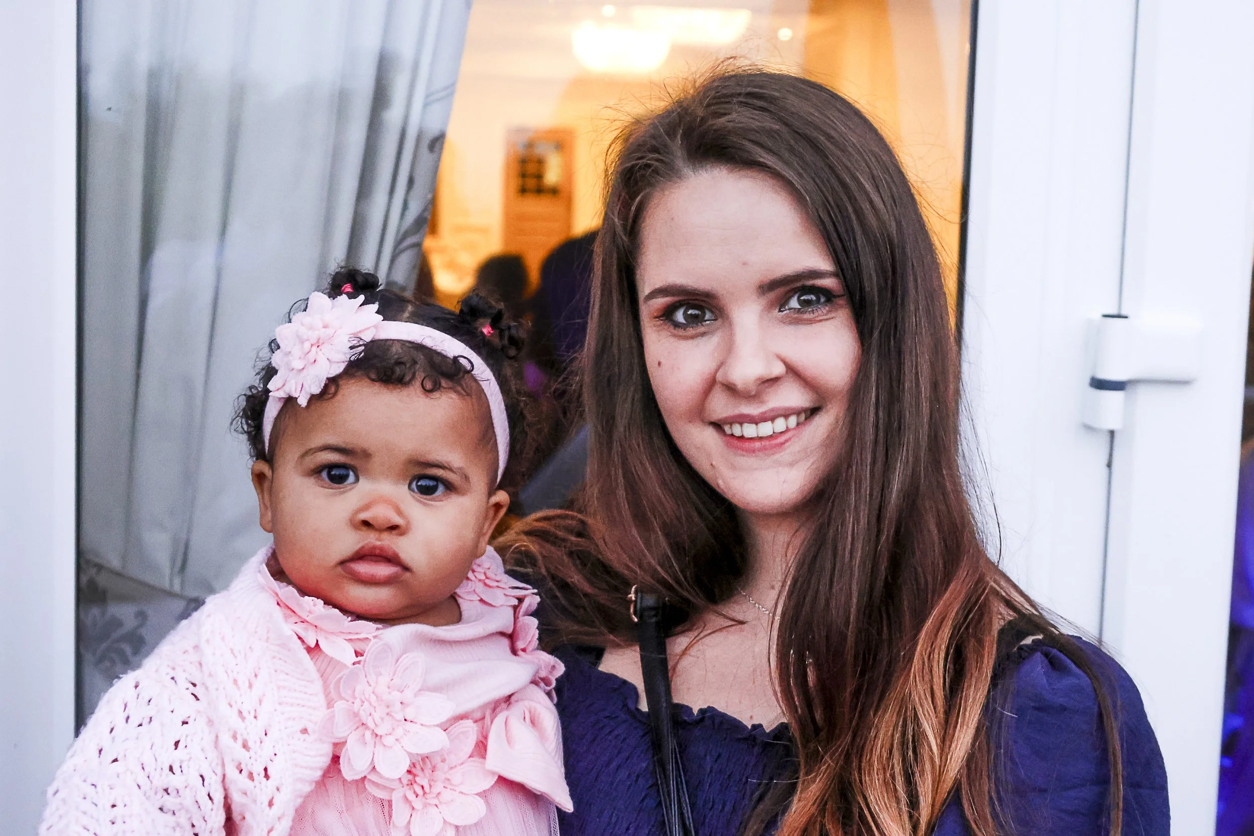A young woman with long brown hair and a young girl with curly hair wearing a pink headband and pink knitted sweater are posing together indoors, possibly at a social event or party. The young woman is smiling, and the girl looks curious, with a neut