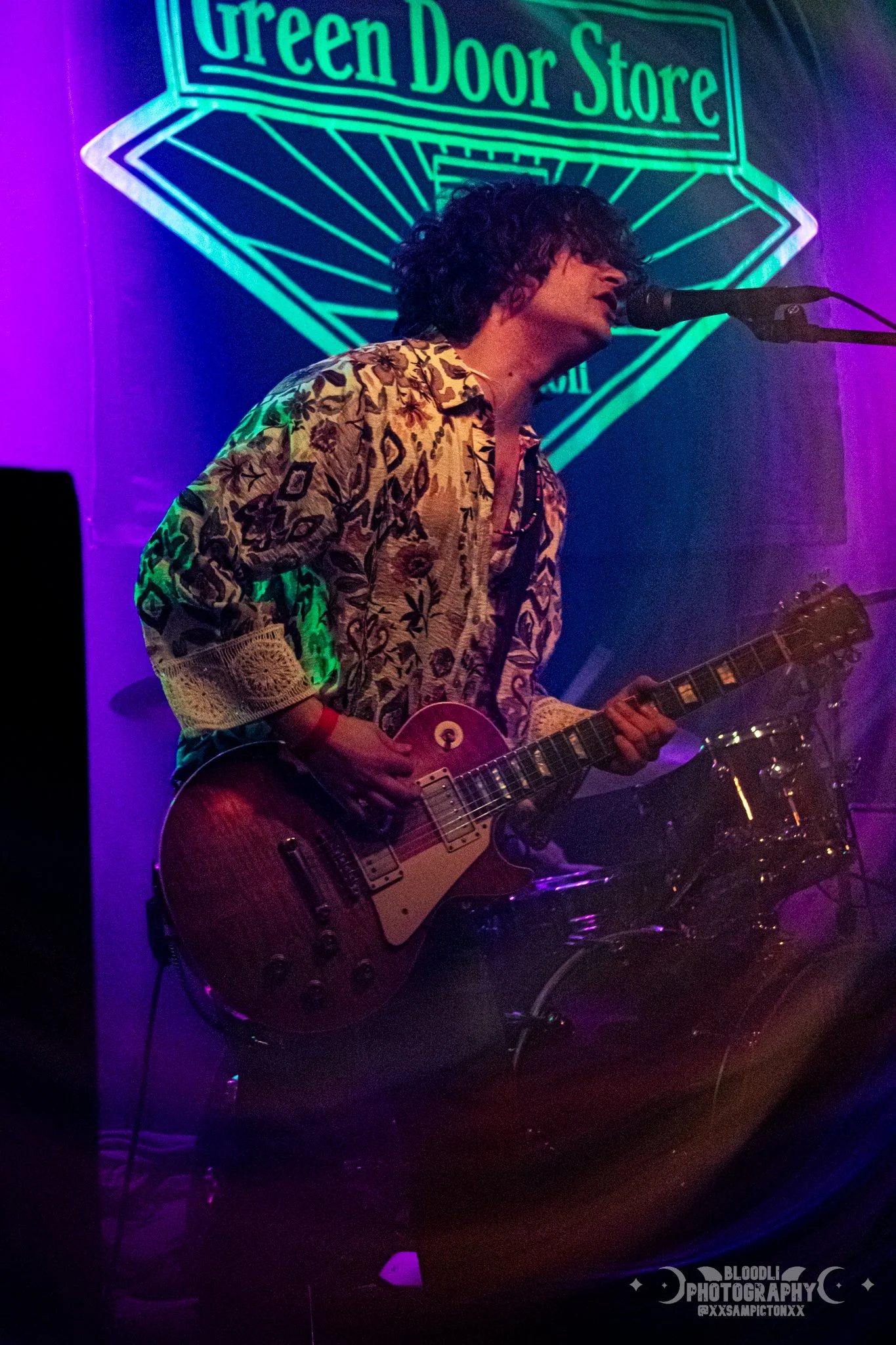 A musician with curly hair, wearing a patterned shirt, playing an electric guitar and singing into a microphone at a live music venue. A neon sign in the background reads 'Green Door Store.'