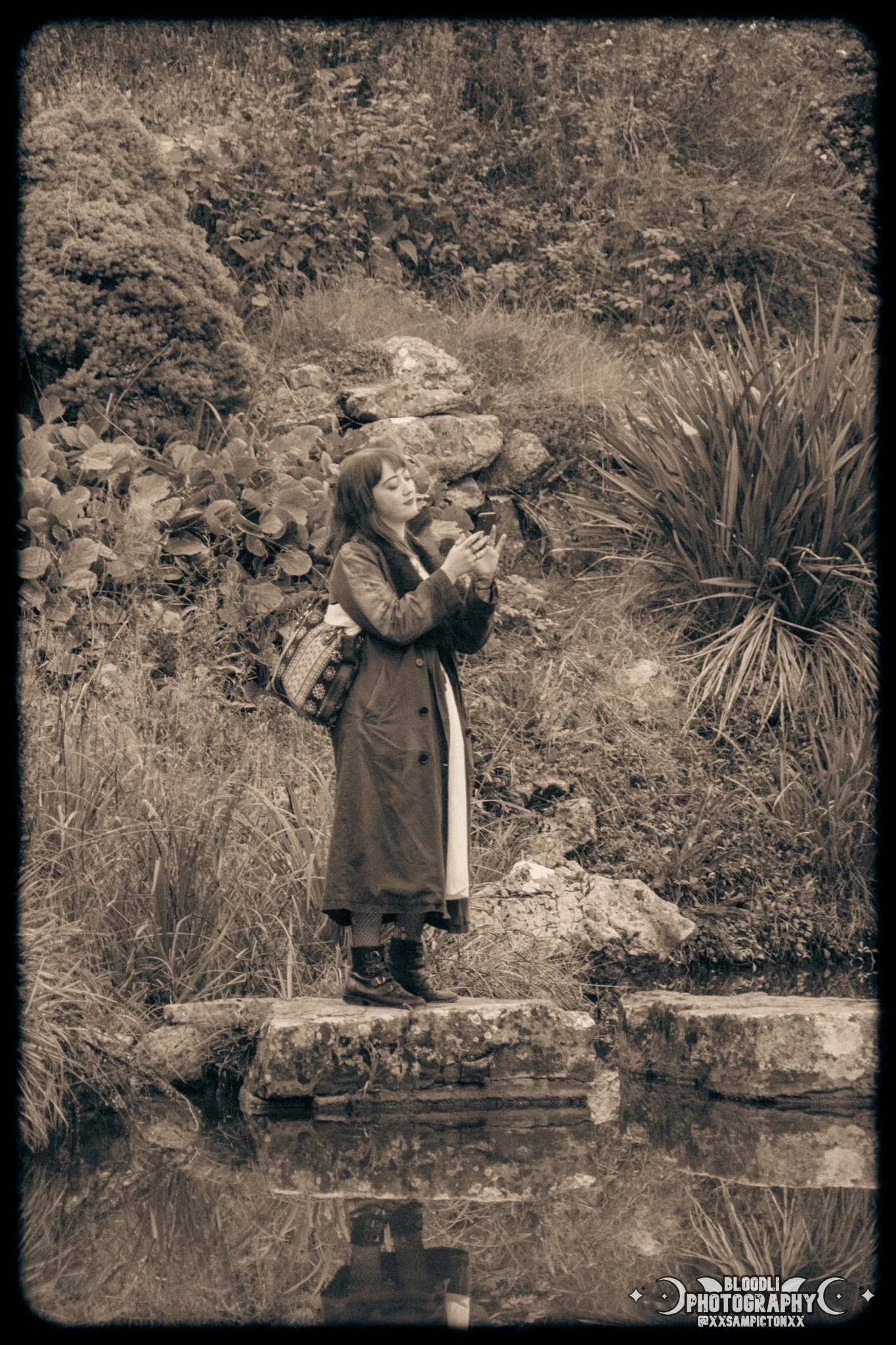 A woman standing on a rock by a pond, using her phone, with lush greenery in the background.
