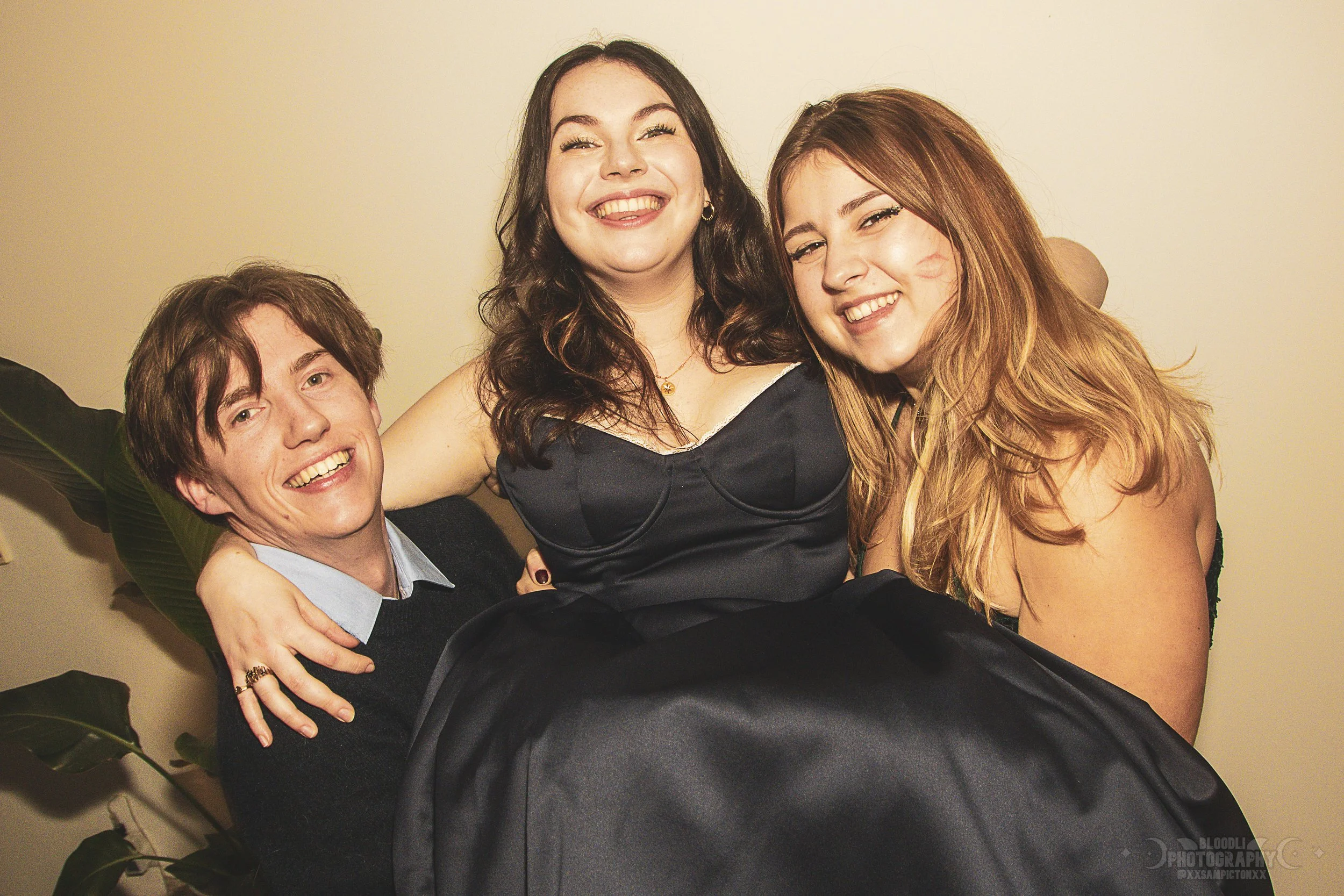 Three young adults smiling and posing for a photo. One man and two women, with the woman in the middle being lifted or supported by the man and the other woman. They are standing indoors against a plain light-colored wall.