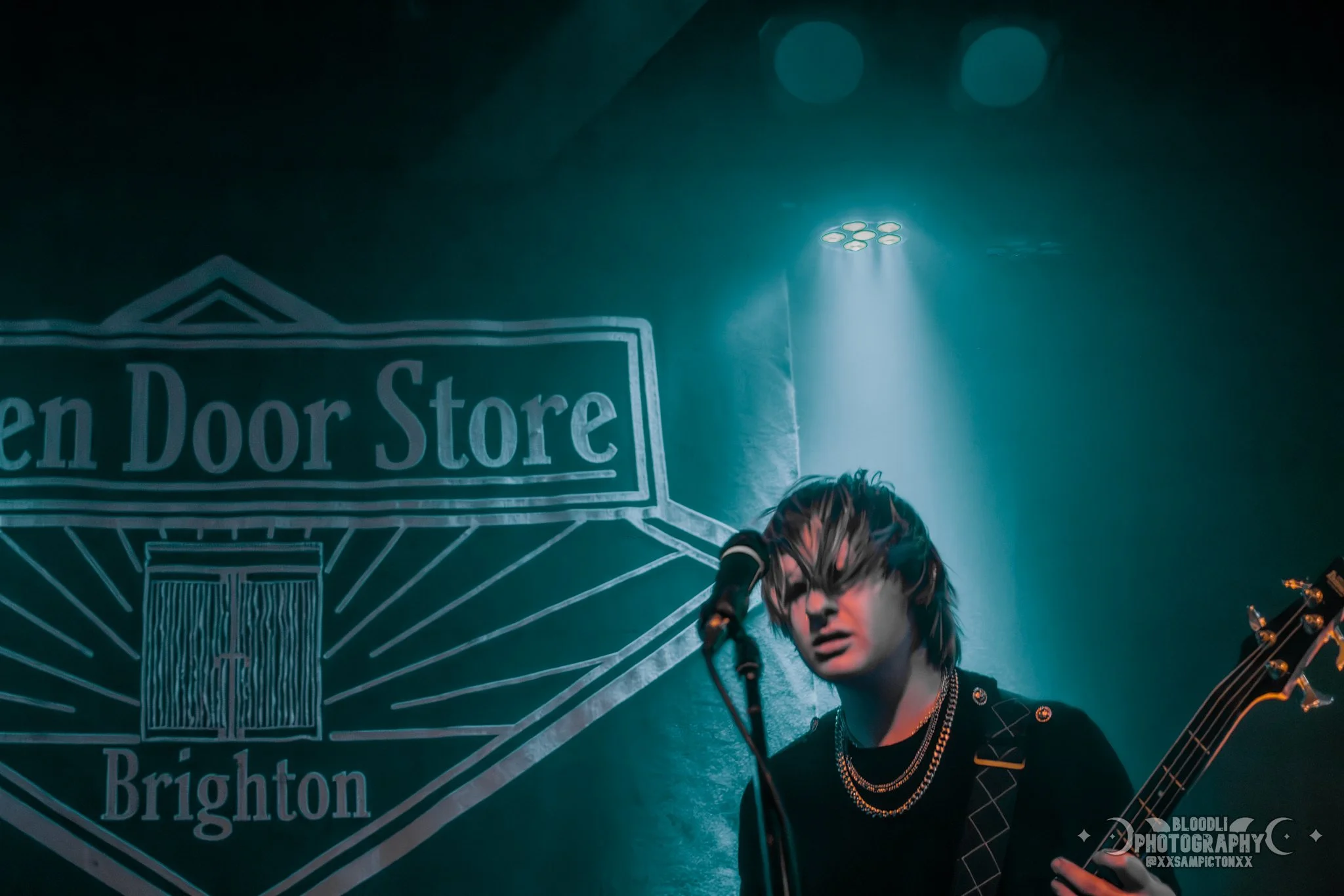 A young musician with shaggy hair singing into a microphone and holding a guitar, illuminated by stage lights, with a sign that reads 'Open Door Store Brighton' in the background.