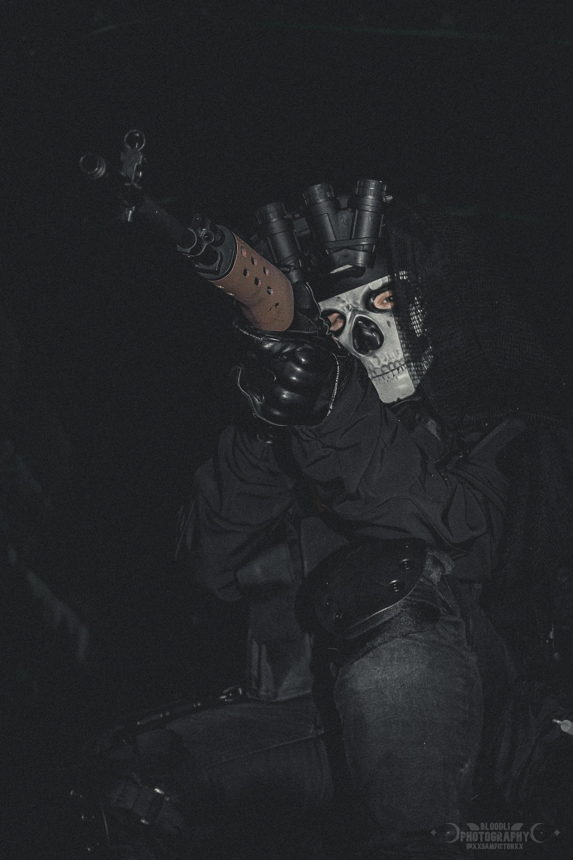 A person wearing a dark hooded jacket and gloves, holding a rifle and aiming. They are wearing a mask resembling a skull with dark eye sockets and nose. The background is black and dimly lit.