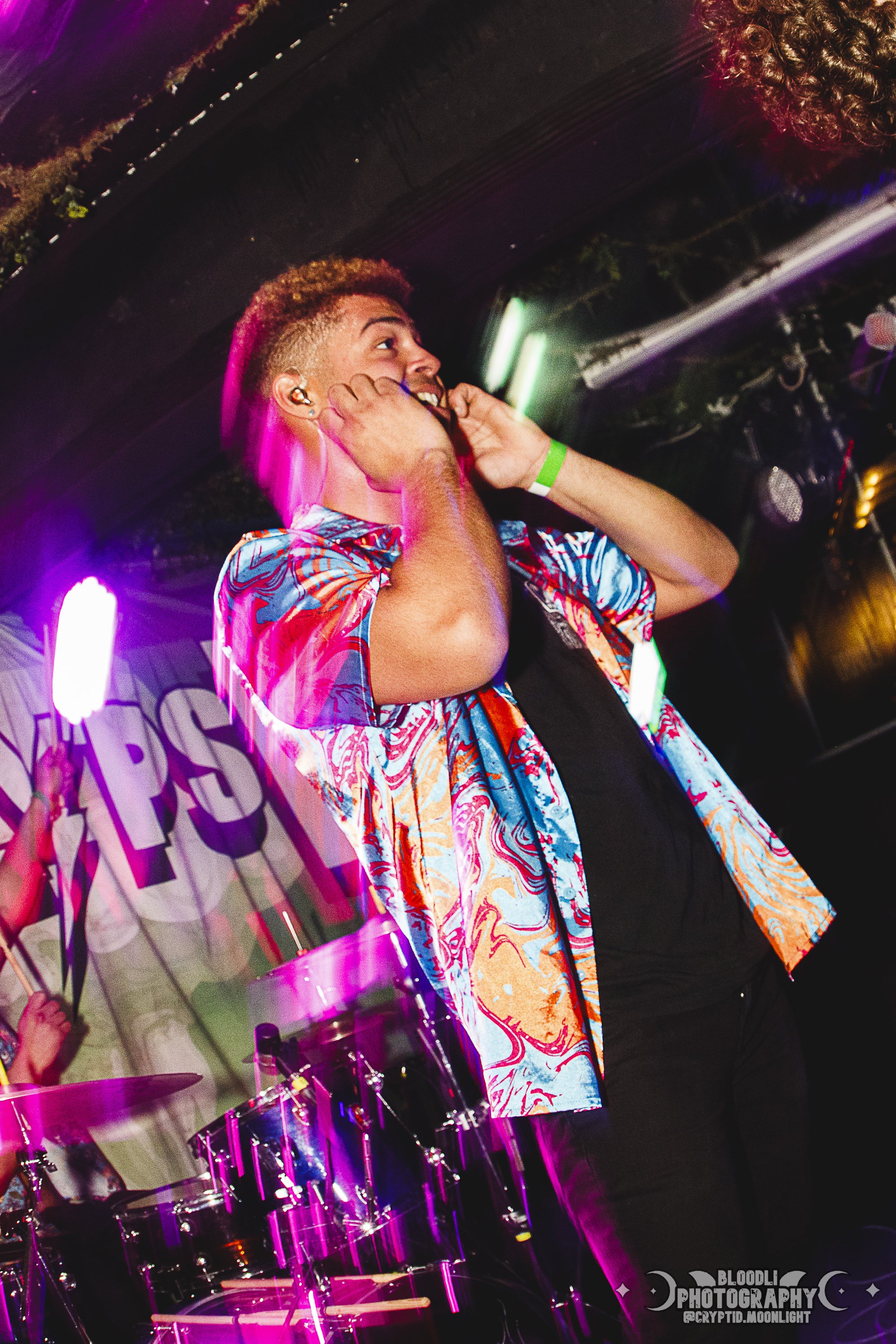A young man with curly hair adjusting his earpiece on stage, colorful lighting, a drum set in the foreground, and a banner with the word 'POSS' partially visible in the background.