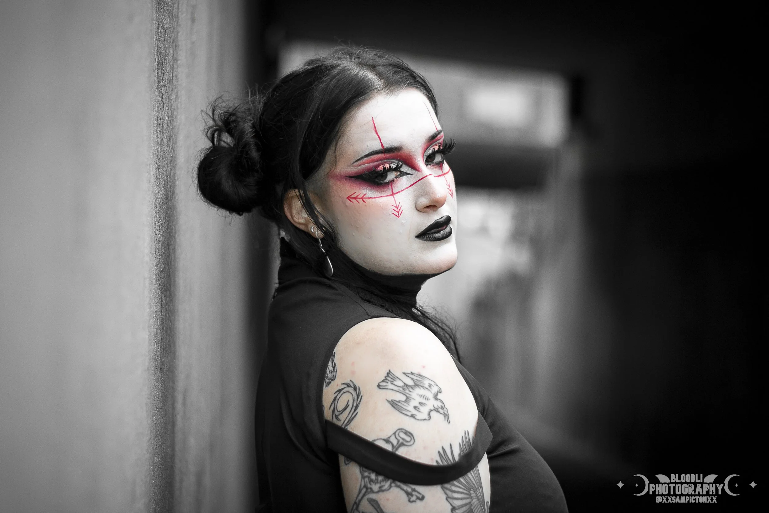 A woman with gothic makeup and tattoos, wearing dark clothing, leaning against a wall in a dimly lit environment.