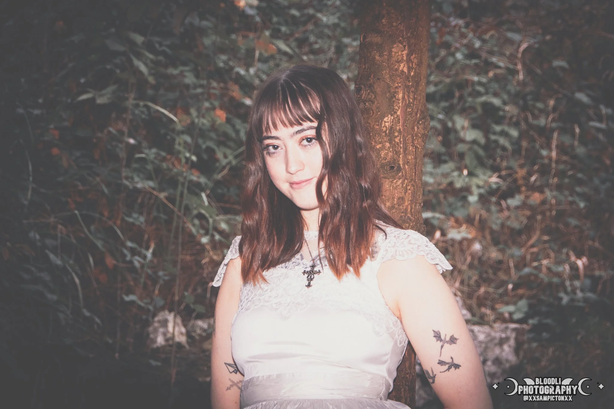 A young woman with wavy brown hair and bangs standing outdoors near a tree in a wooded area, wearing a white dress with lace details and a necklace with a cross pendant, with tattoos on her arms.
