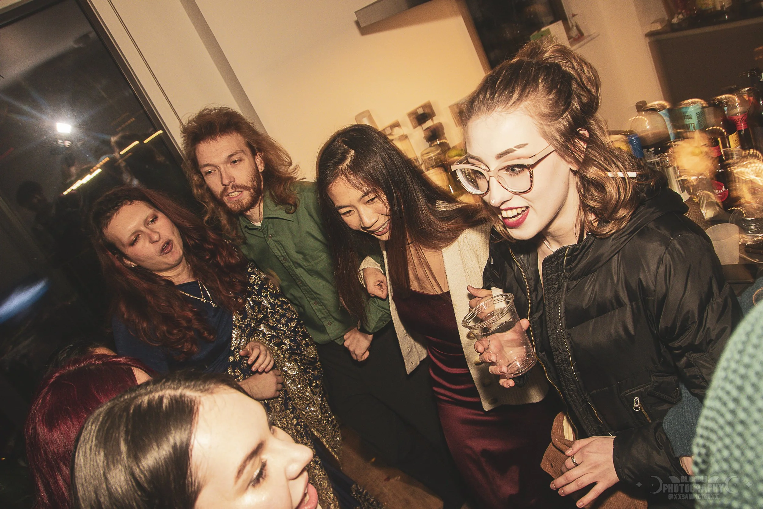 Group of five people at a social gathering, standing close together, smiling and laughing, with a bar or snack table and jars in the background.