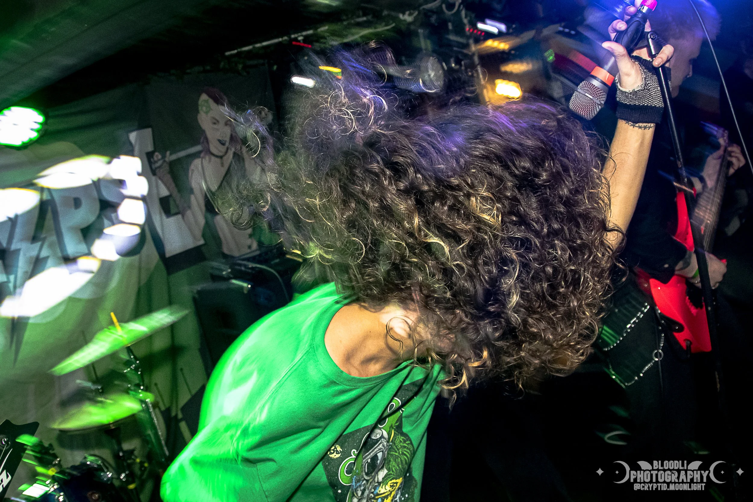 A person with long curly hair wearing a green T-shirt playing drums at a music event. The background features a banner and musical equipment, with vibrant colorful lighting.
