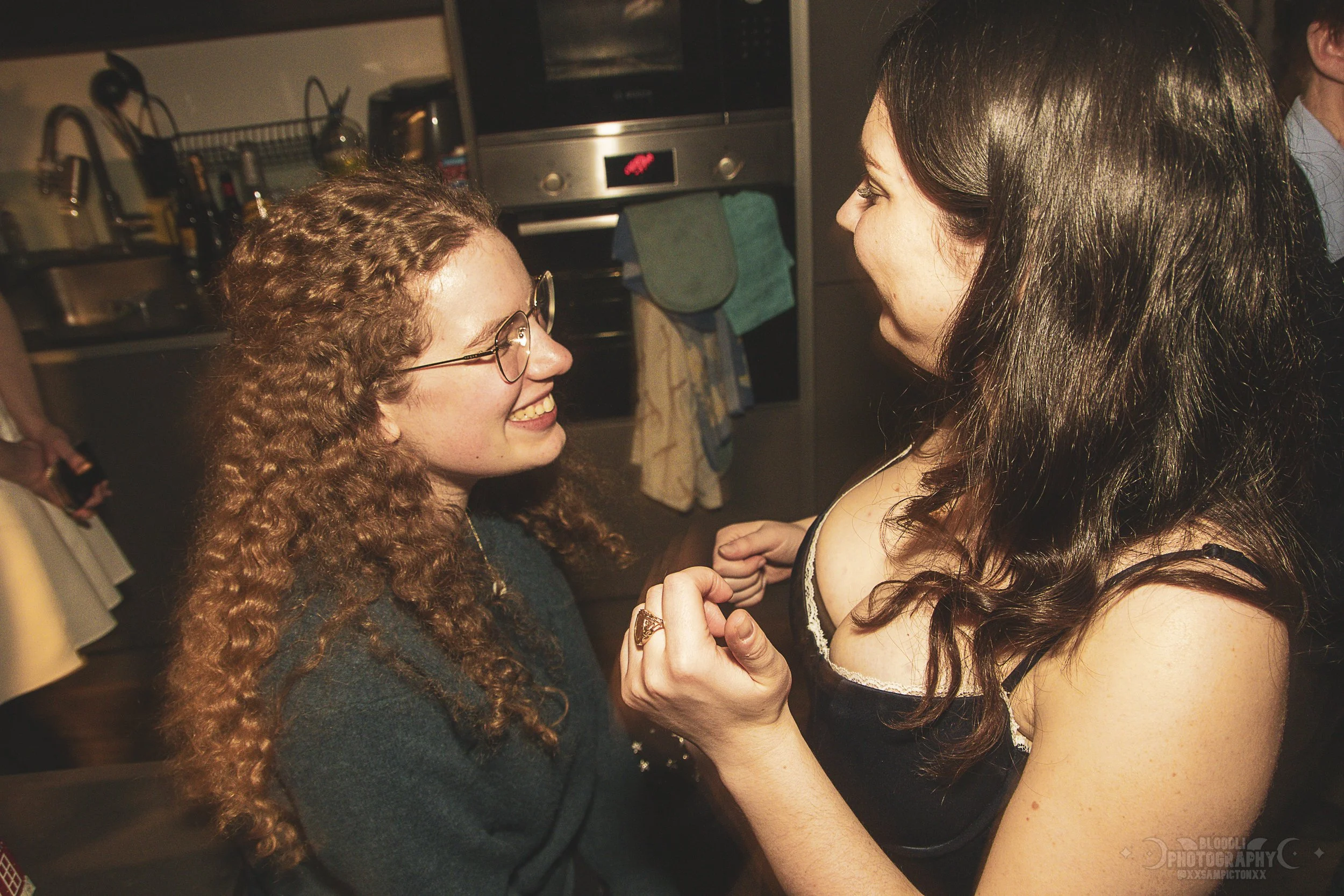 Two women are smiling and holding hands in a kitchen, engaging in a close conversation or moment. One has long, curly hair and glasses, the other has long, straight dark hair.