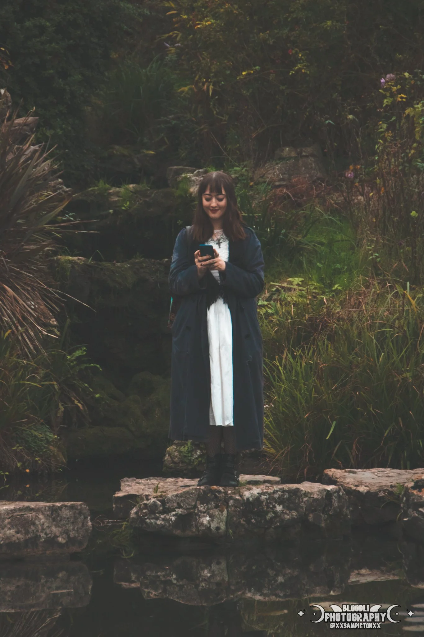 A young woman standing on a stone pathway next to a small pond, surrounded by lush green vegetation, while looking at her smartphone.