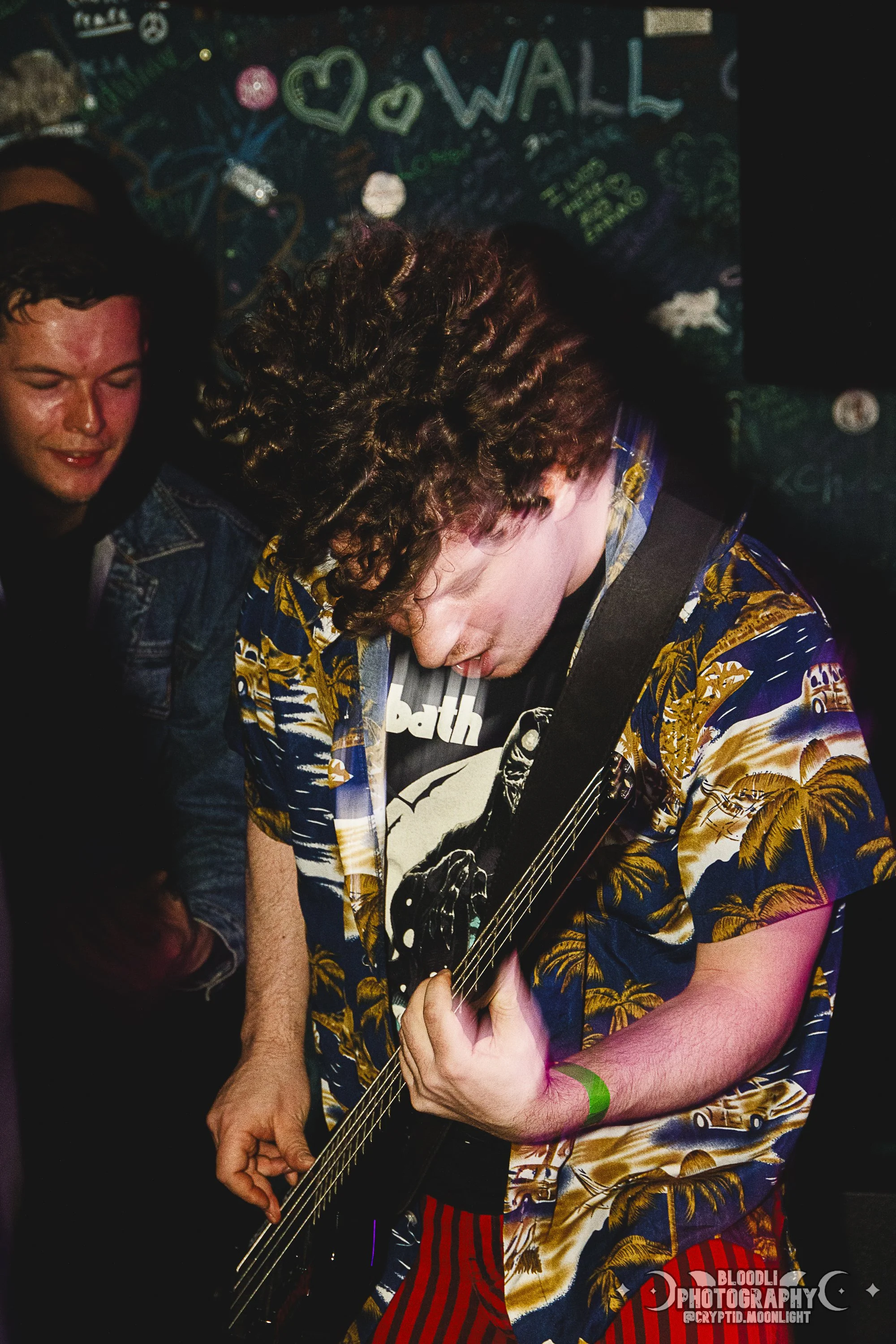 A young man with curly hair playing an electric guitar at a music venue, wearing a colorful Hawaiian shirt and a black graphic t-shirt, with people and a chalkboard wall in the background.