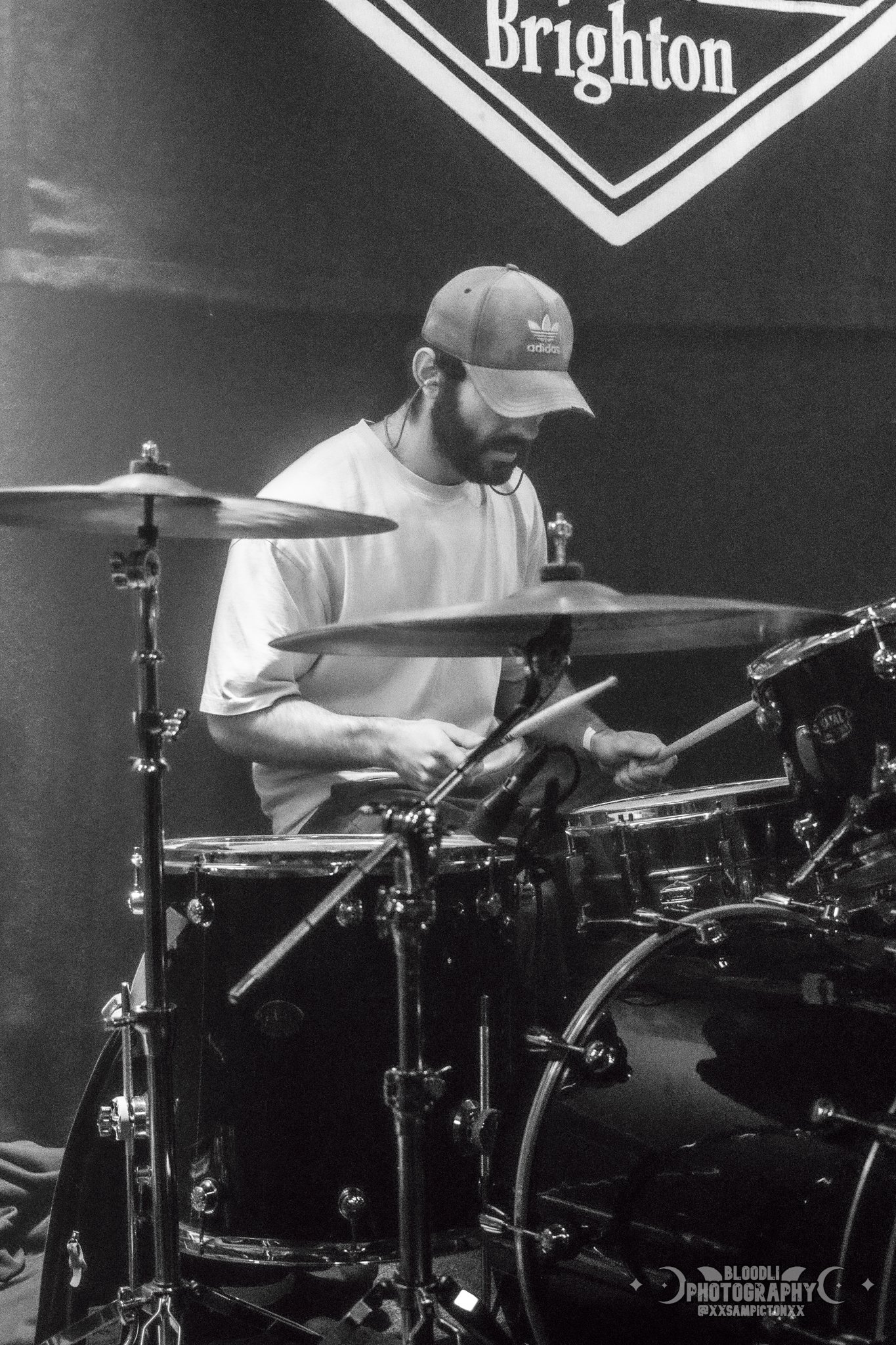 A man playing a drum set on stage, wearing a baseball cap and a white shirt, with a Brighton sign in the background.