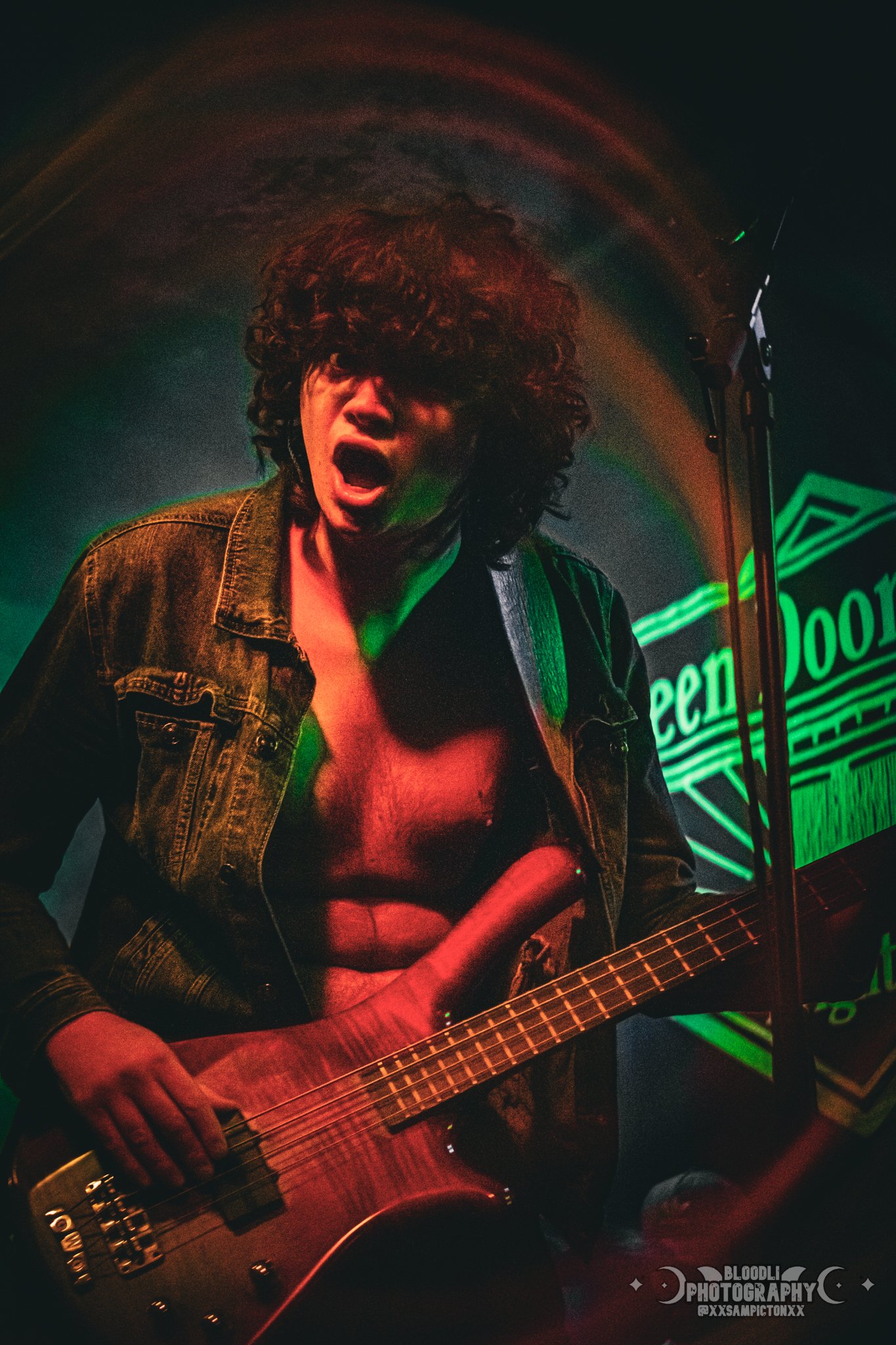A person with curly hair playing an electric bass guitar on stage, with a neon green sign that says 'Open Door' in the background, wearing a denim jacket, no shirt underneath, and singing or shouting passionately.