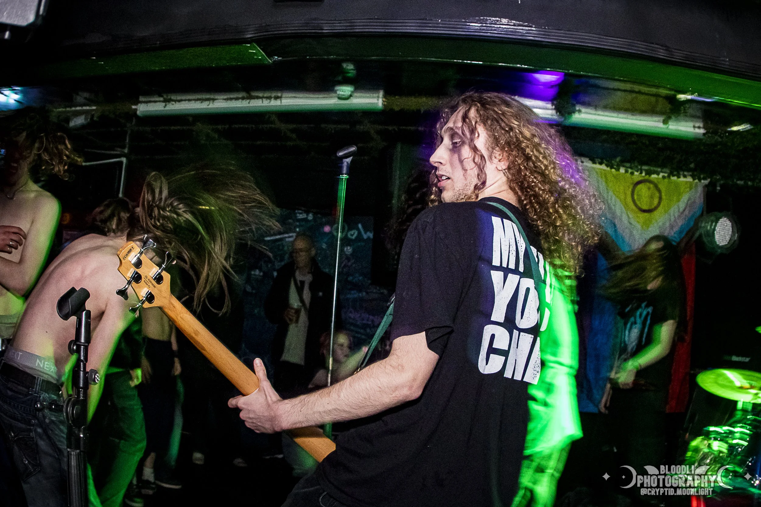 A person with long, curly hair playing guitar in a nightclub with colorful lighting, surrounded by people dancing.