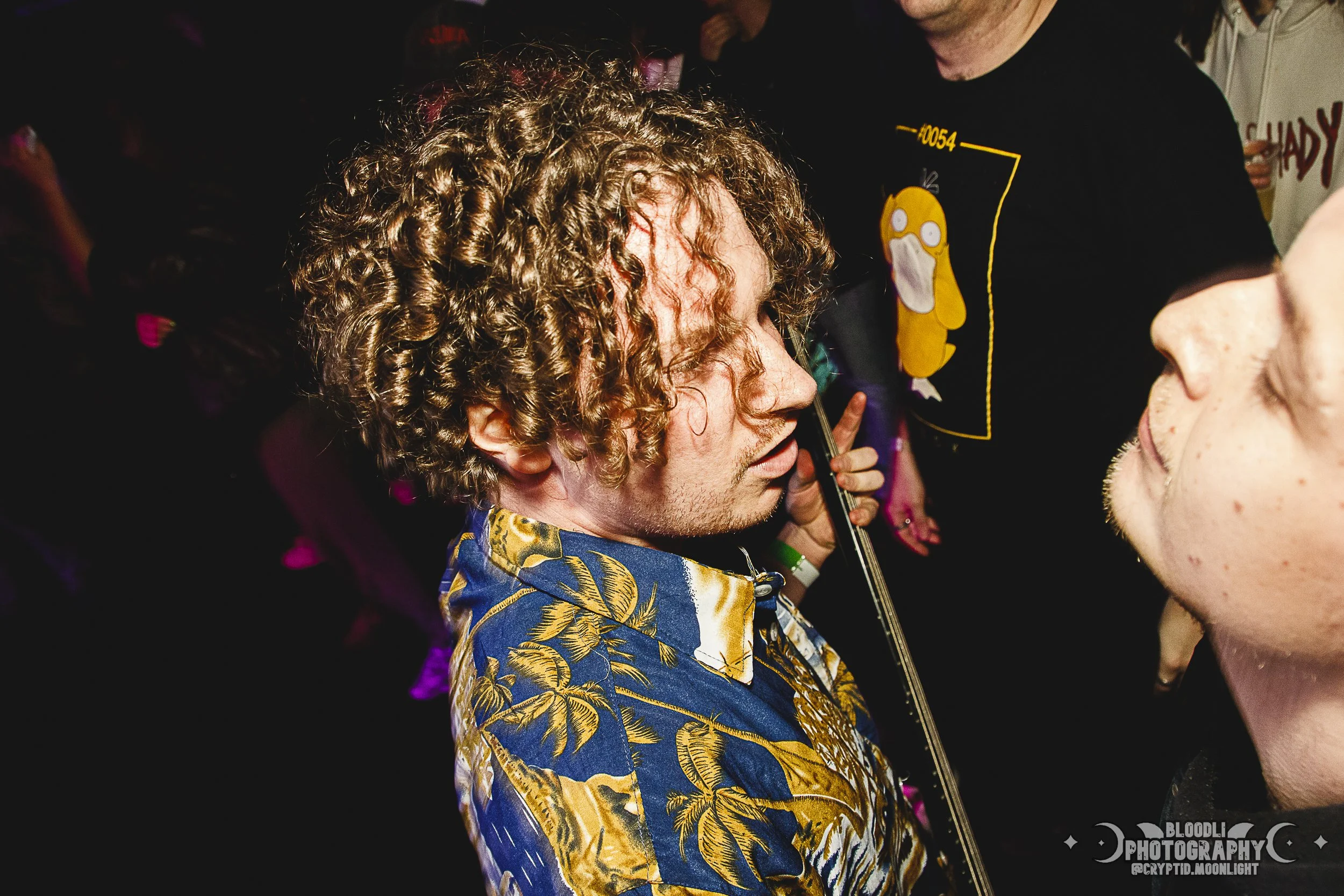 A man with curly hair playing a violin at a party or concert, wearing a colorful Hawaiian shirt, with people in the background.