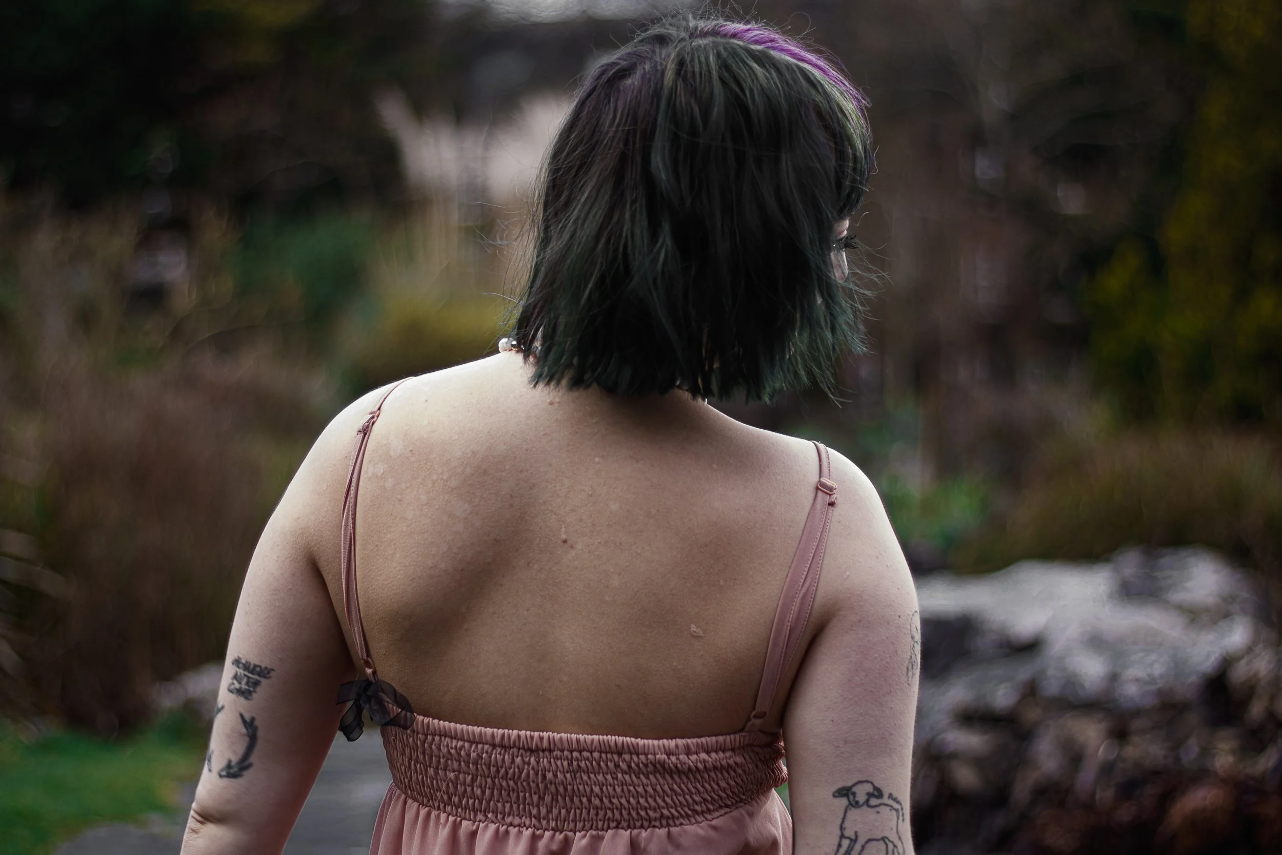 Back view of a woman with chin-length black hair with purple and green tips, wearing a pink spaghetti strap dress, outdoors in a natural setting with blurred background.