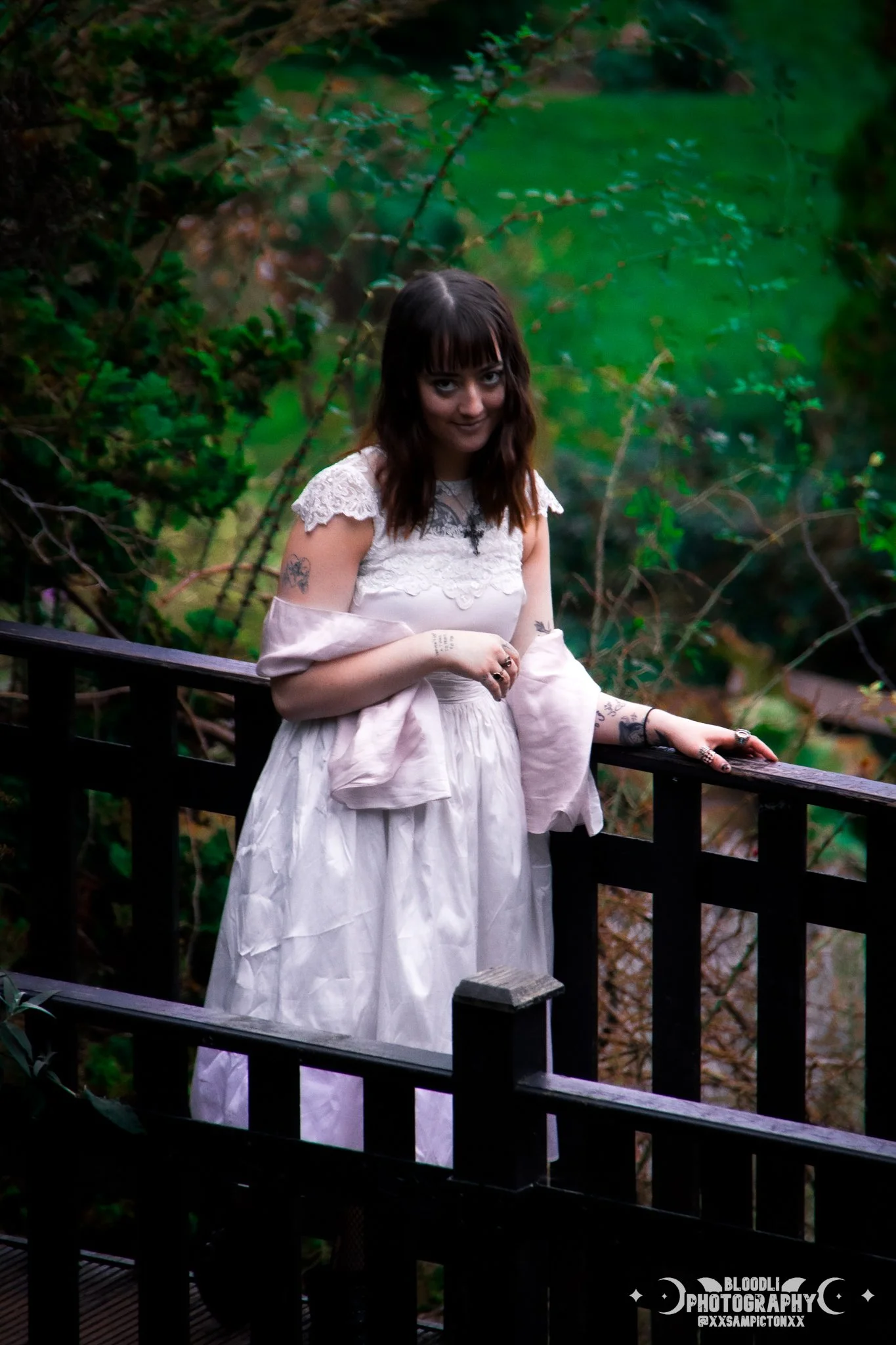 A young woman with dark hair and tattoos on her arms, wearing a white dress, standing on a wooden bridge in a lush green forest, looking at the camera.