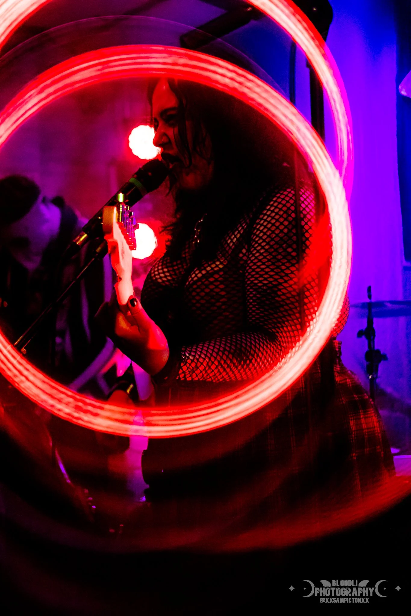 A woman wearing fishnet clothing singing into a microphone at a concert, with colorful, circular light trails around her.