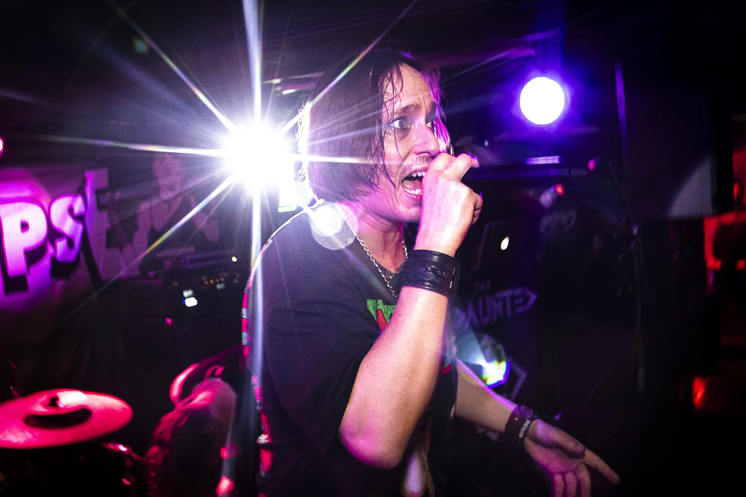 A performer singing into a microphone on stage with bright colorful lights shining behind them, wearing black clothing and accessories.