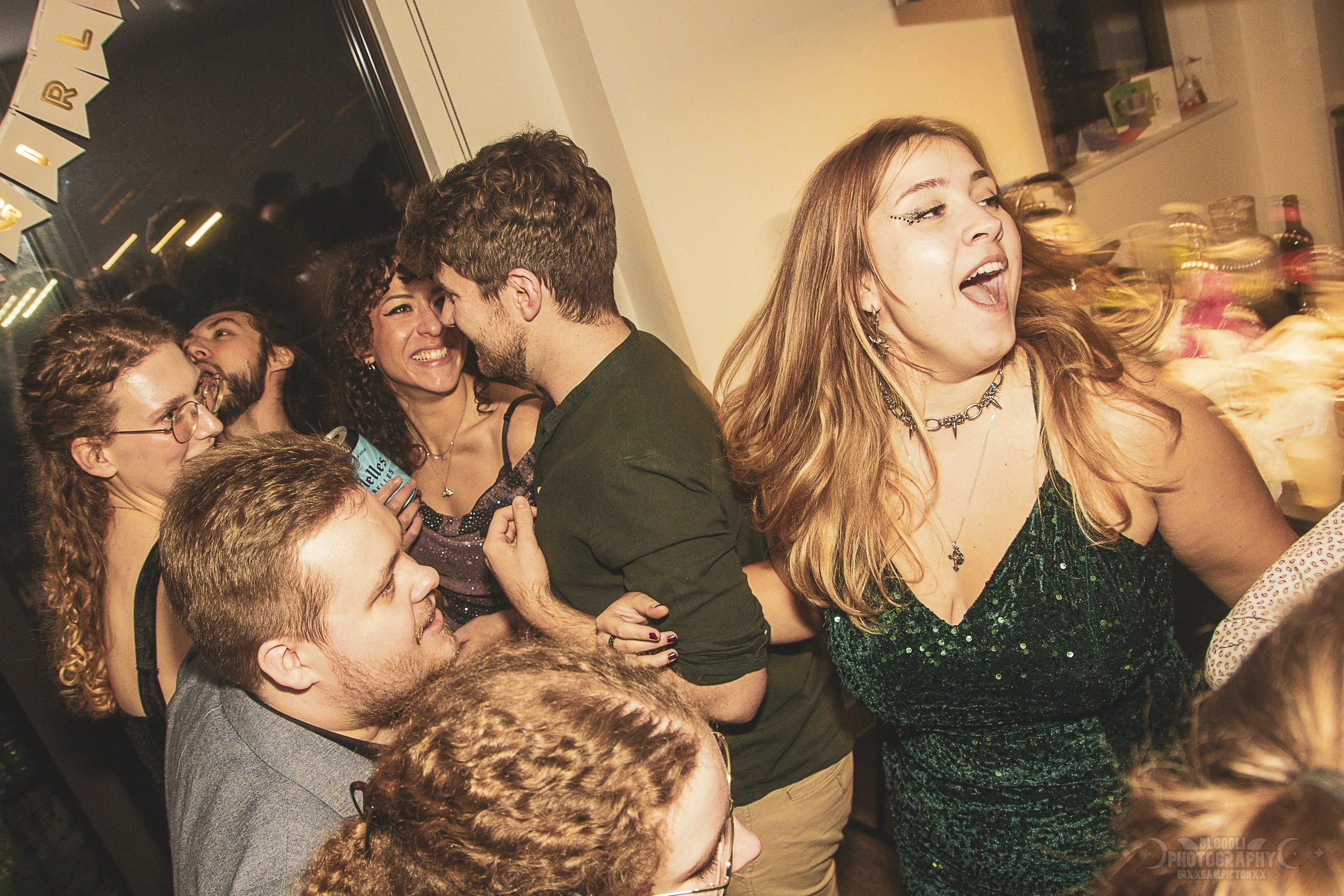Group of young adults at a party, dancing and smiling with a woman in a green dress singing or talking loudly, others laughing and enjoying themselves.