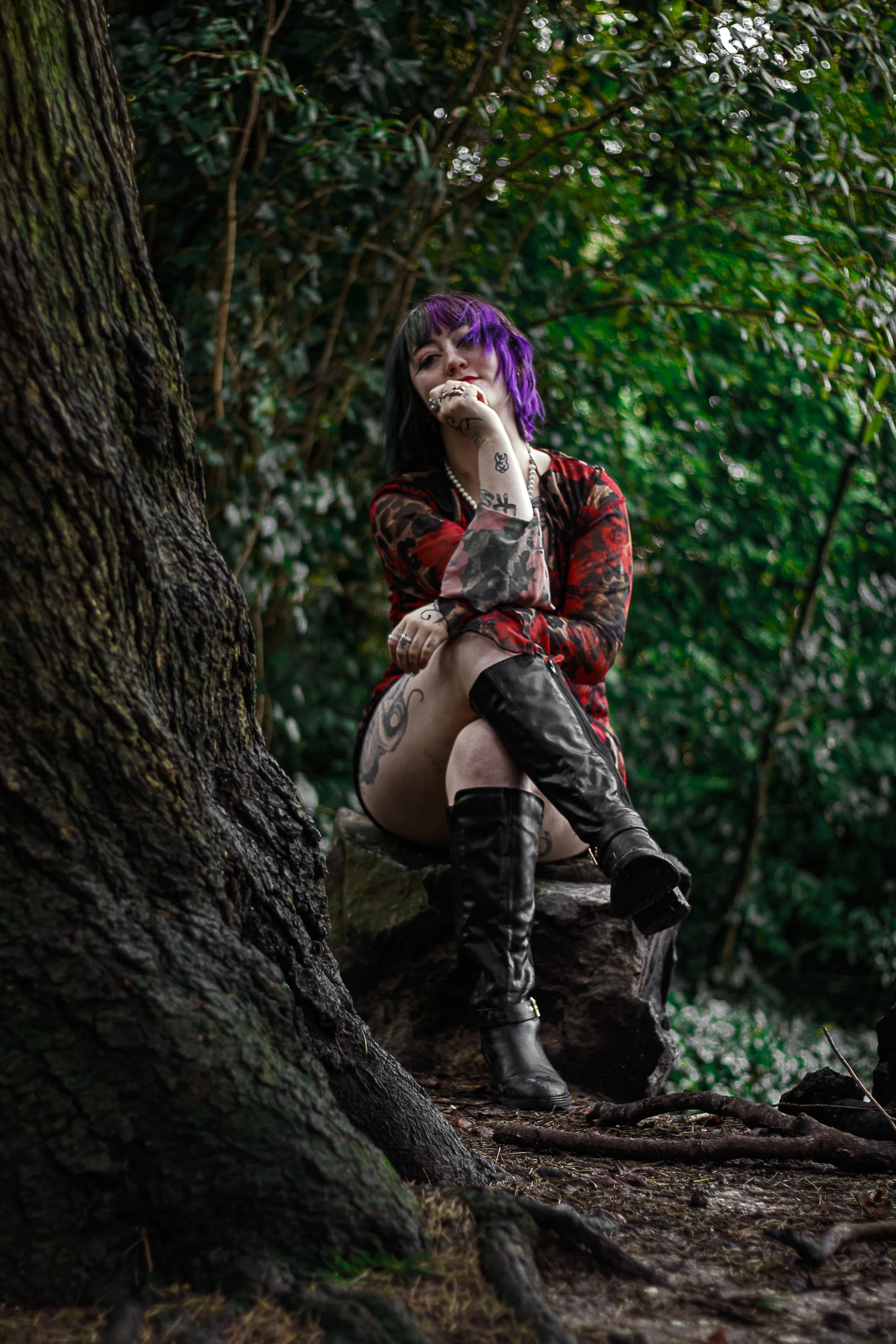 A woman with purple and black hair, wearing a red and black dress with tattoos, black thigh-high boots, sitting on a rock in a wooded area.