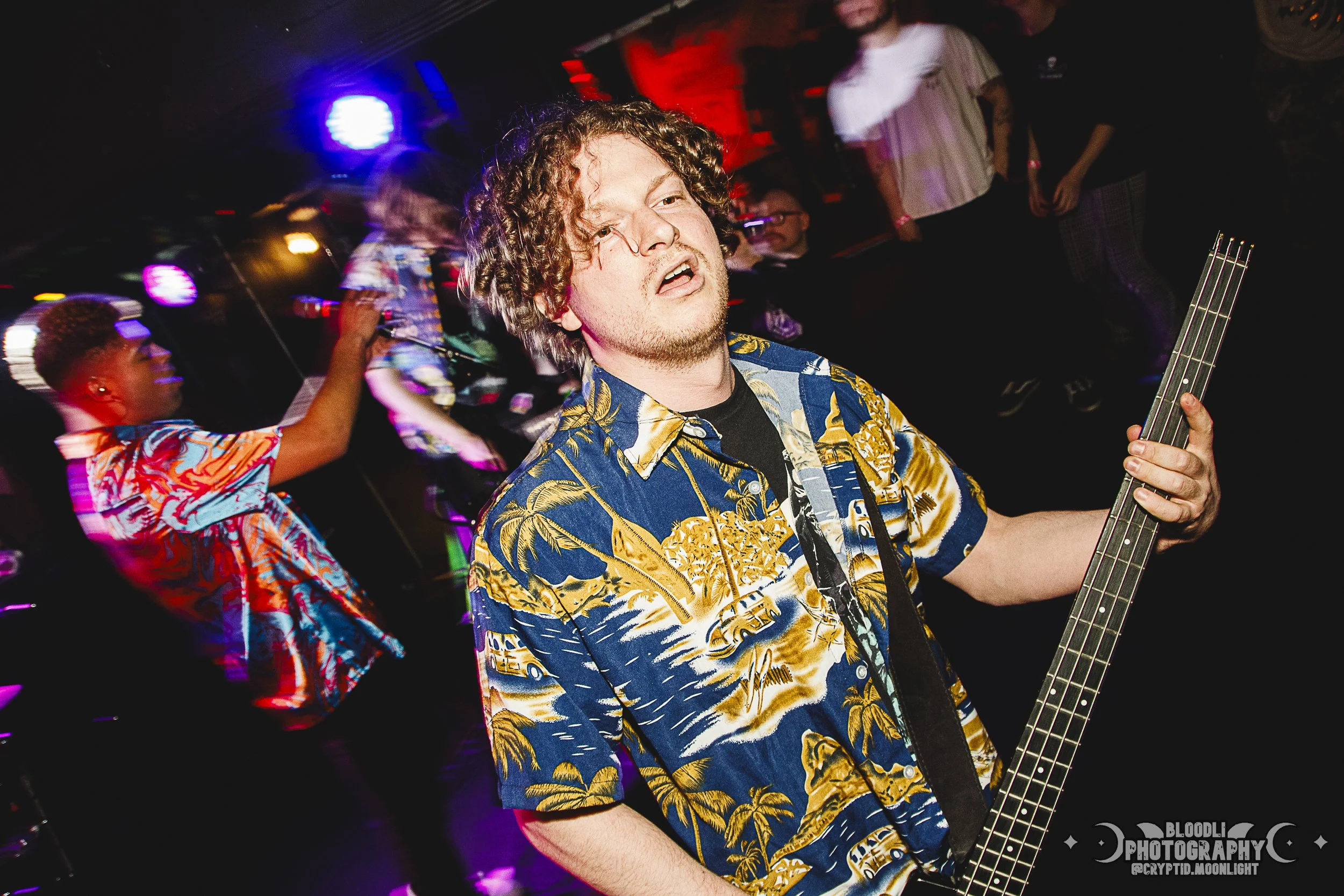 A man with curly hair wearing a Hawaiian shirt is playing an electric guitar in a nightclub. In the background, people are dancing with colorful lighting and blurred motion.