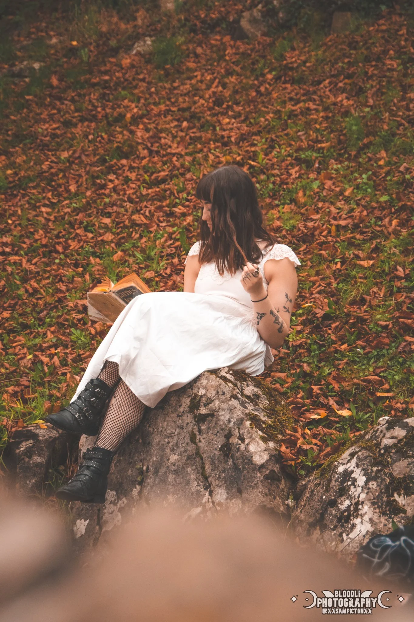 A woman with tattoos on her arm and dark hair in a bob cut, wearing a white dress and fishnet stockings, sitting on a rock in a forest with fallen leaves, reading a book.