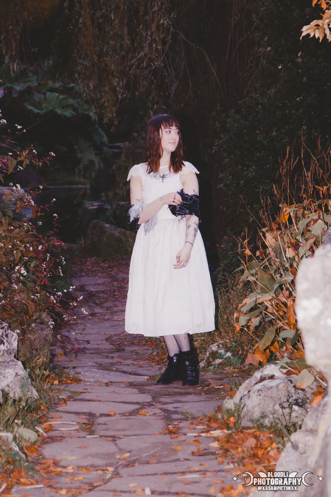 A woman with tattoos, wearing a white dress, black boots, and fishnet stockings, standing on a stone pathway surrounded by fall foliage and trees at night.