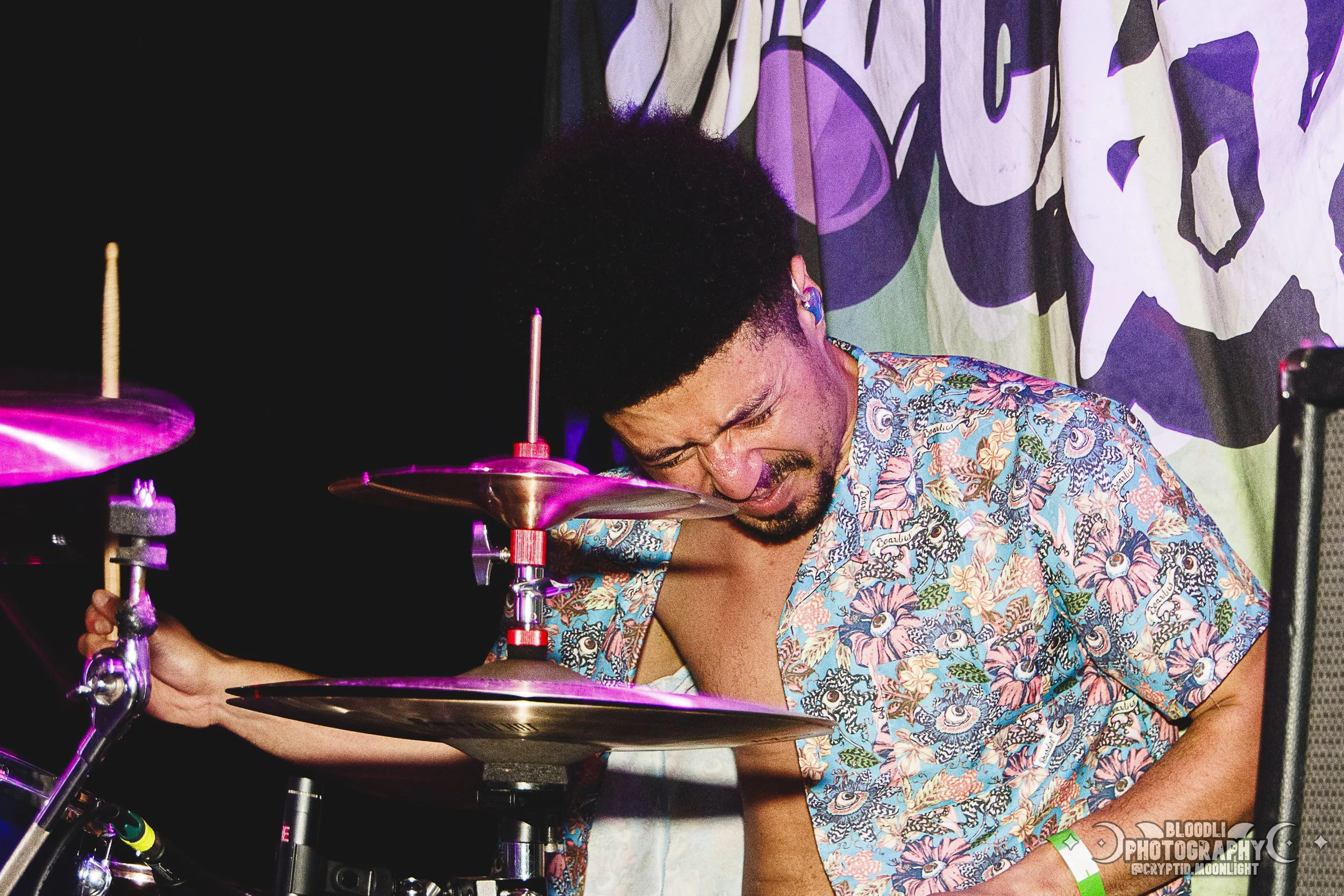 A man with a curly afro hairstyle, wearing a colorful Hawaiian shirt, intensely playing drums with a pained or concentrated expression, in a dimly lit setting with a patterned backdrop.