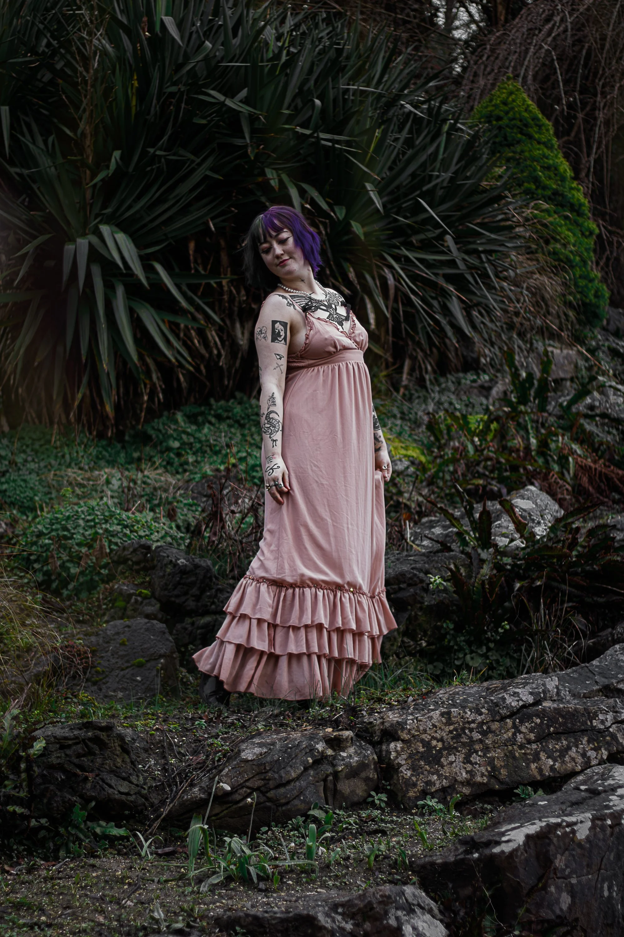 Young woman with purple and black hair standing outdoors on rocks, wearing a pink dress with ruffled hem, surrounded by lush green plants and rocks.