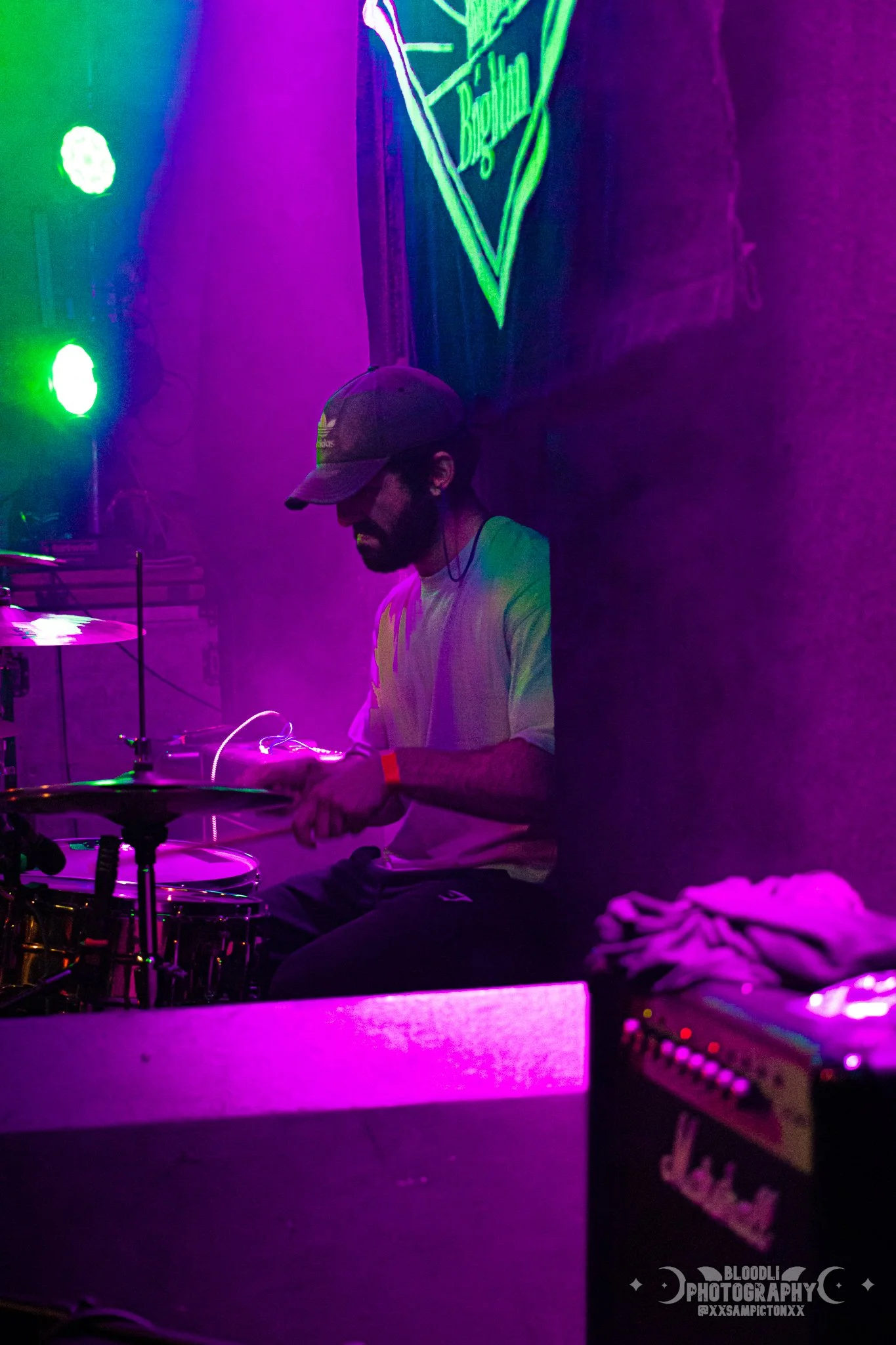 A man wearing a gray cap and white t-shirt playing drums on a stage with colorful purple and green lighting, and a neon sign in the background.