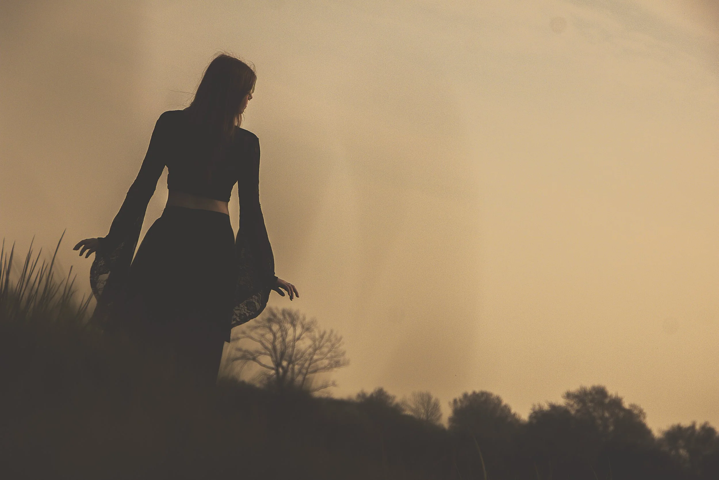 A woman in a black outfit standing outdoors at sunset, with trees in the background.