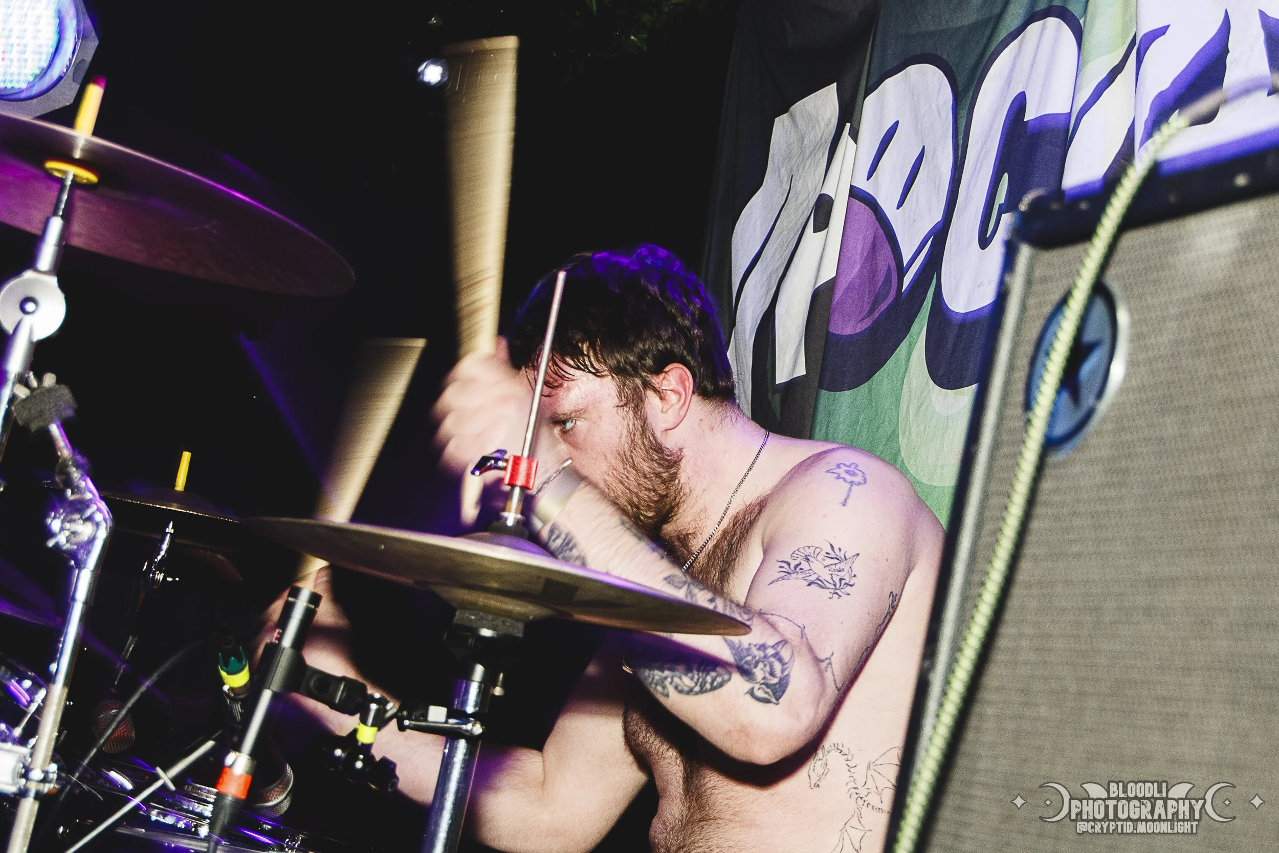 A shirtless man with tattoos on his arm and chest playing drums on stage during a concert.
