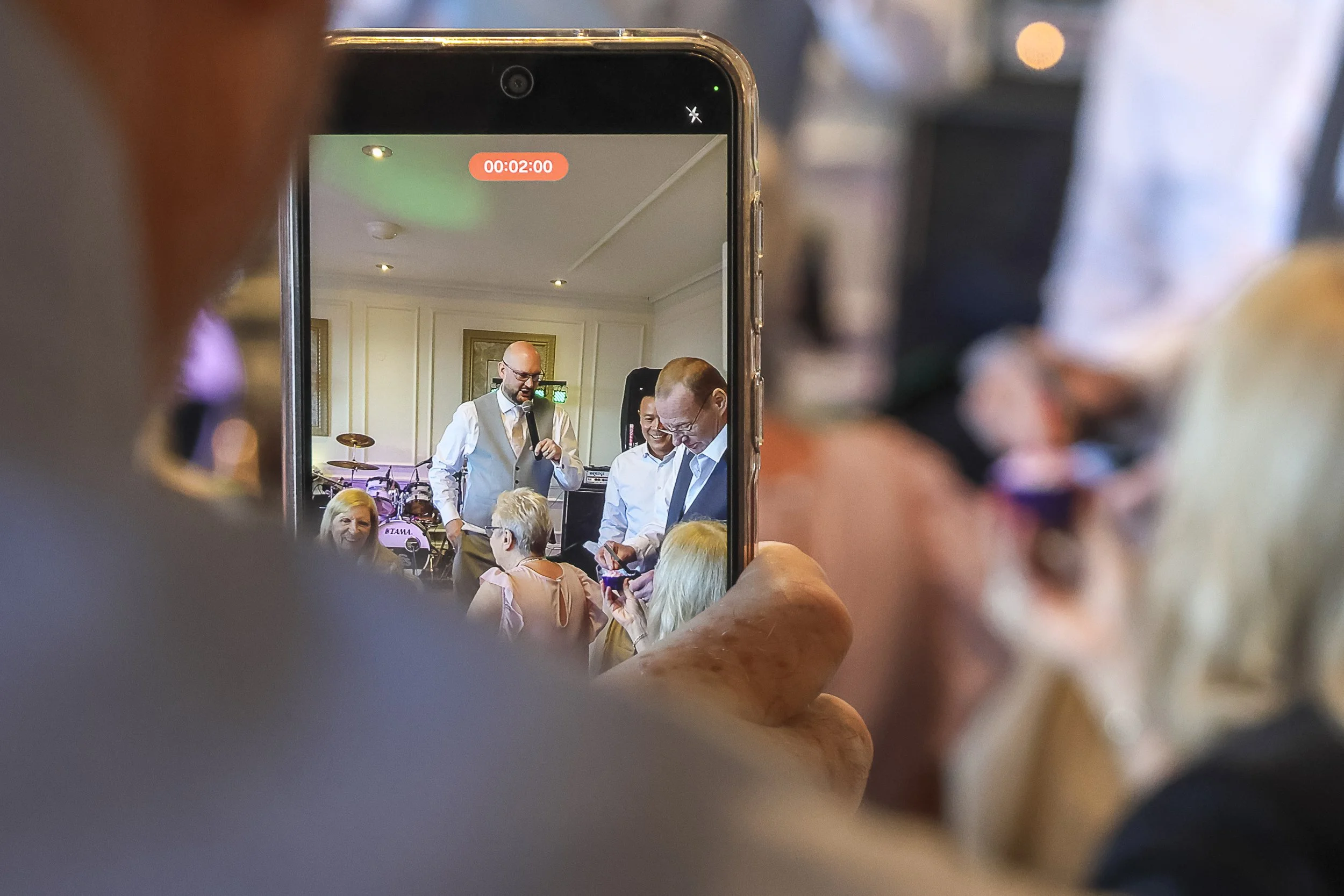 A smartphone captures a group of people at a formal indoor event, with three men standing and speaking while others, including women, listen and take photos. The group appears to be engaged in a discussion or presentation.