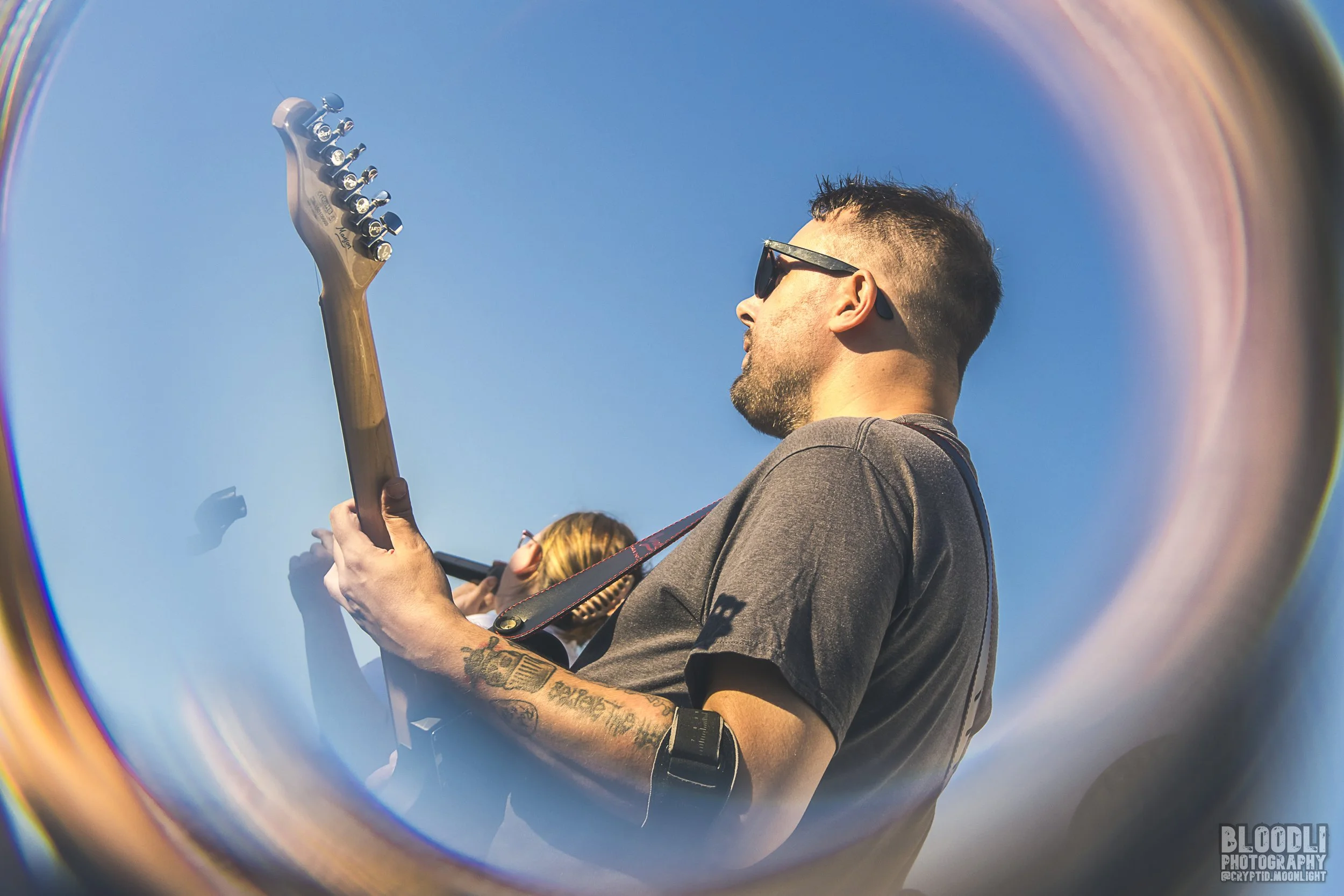 A man playing an electric guitar, wearing sunglasses and a gray shirt, is seen through a circular reflective surface with a clear blue sky in the background.
