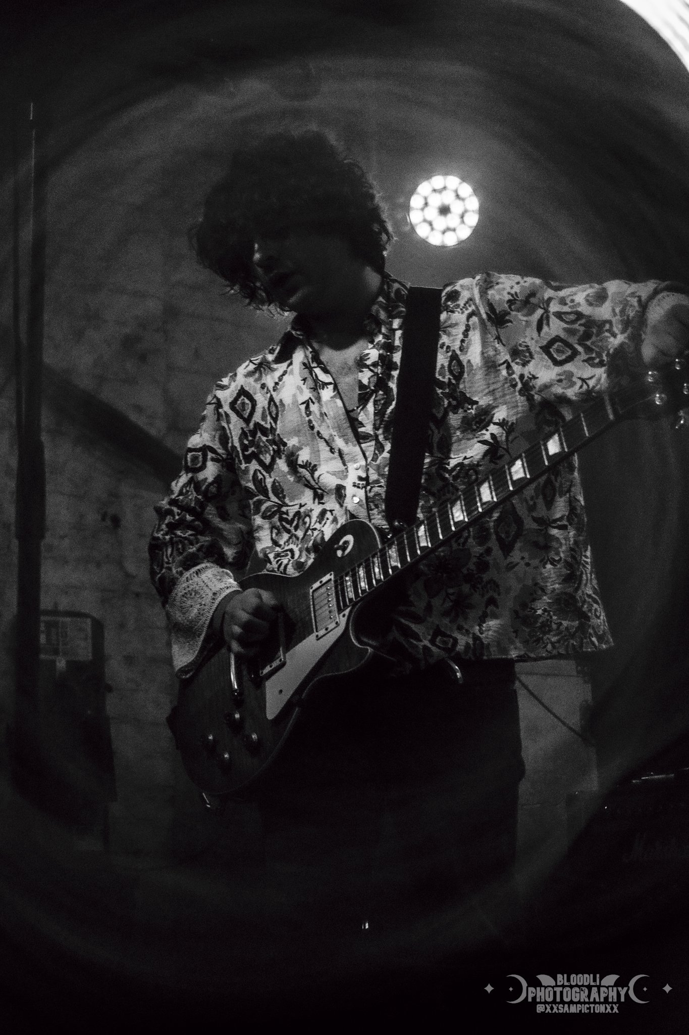 A black and white photo of a person with curly hair wearing a patterned shirt playing an electric guitar indoors with a brick wall in the background.