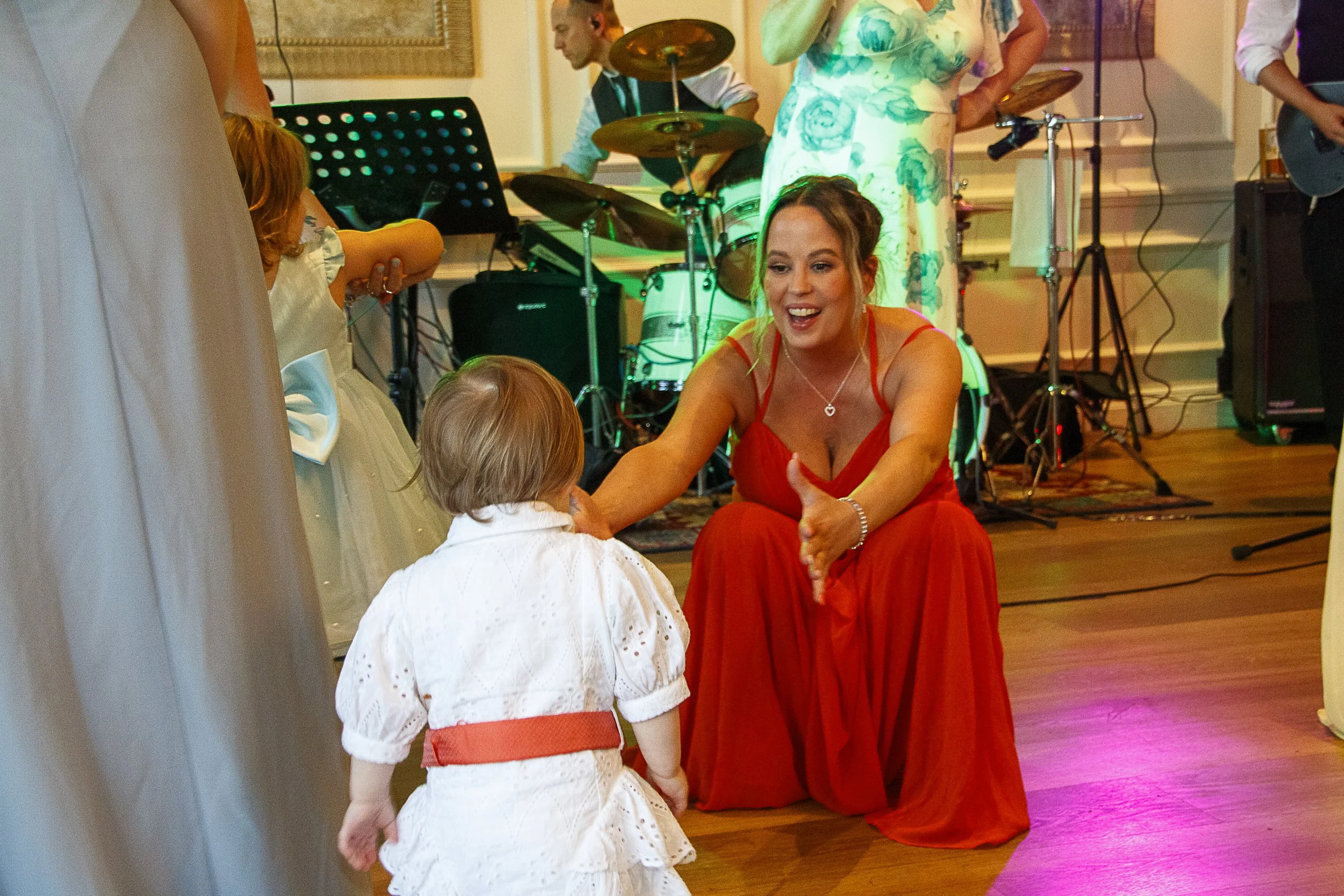A woman in a red dress smiling and reaching out to a young girl in a white dress at a celebration, with a live band playing in the background.