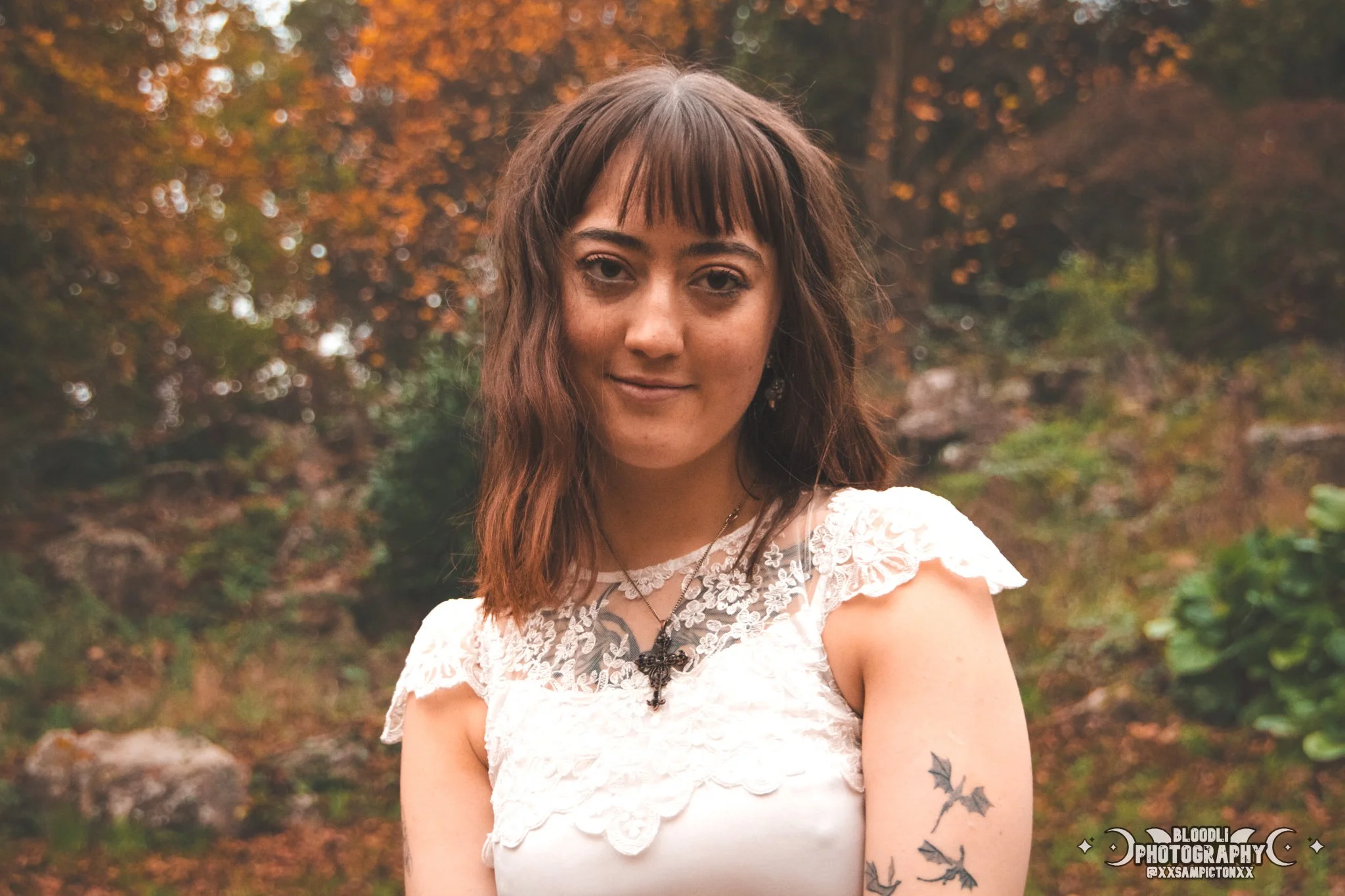 A young woman with medium-length brown hair, wearing a white lace dress and a black cross necklace, stands outdoors in front of autumn trees. She has a tattoo of black bats on her upper right arm and a small smile on her face.
