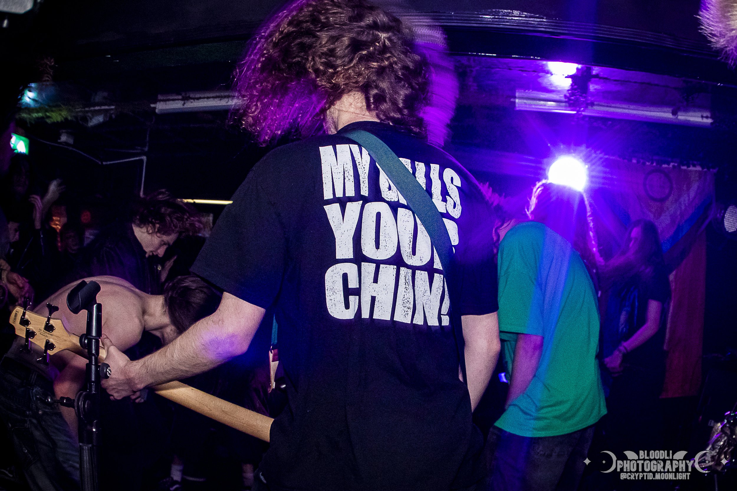 People at a concert or music event, with a person in the center wearing a black shirt that says 'MY GUNS ARE YOUR CHAIN' under purple lighting.