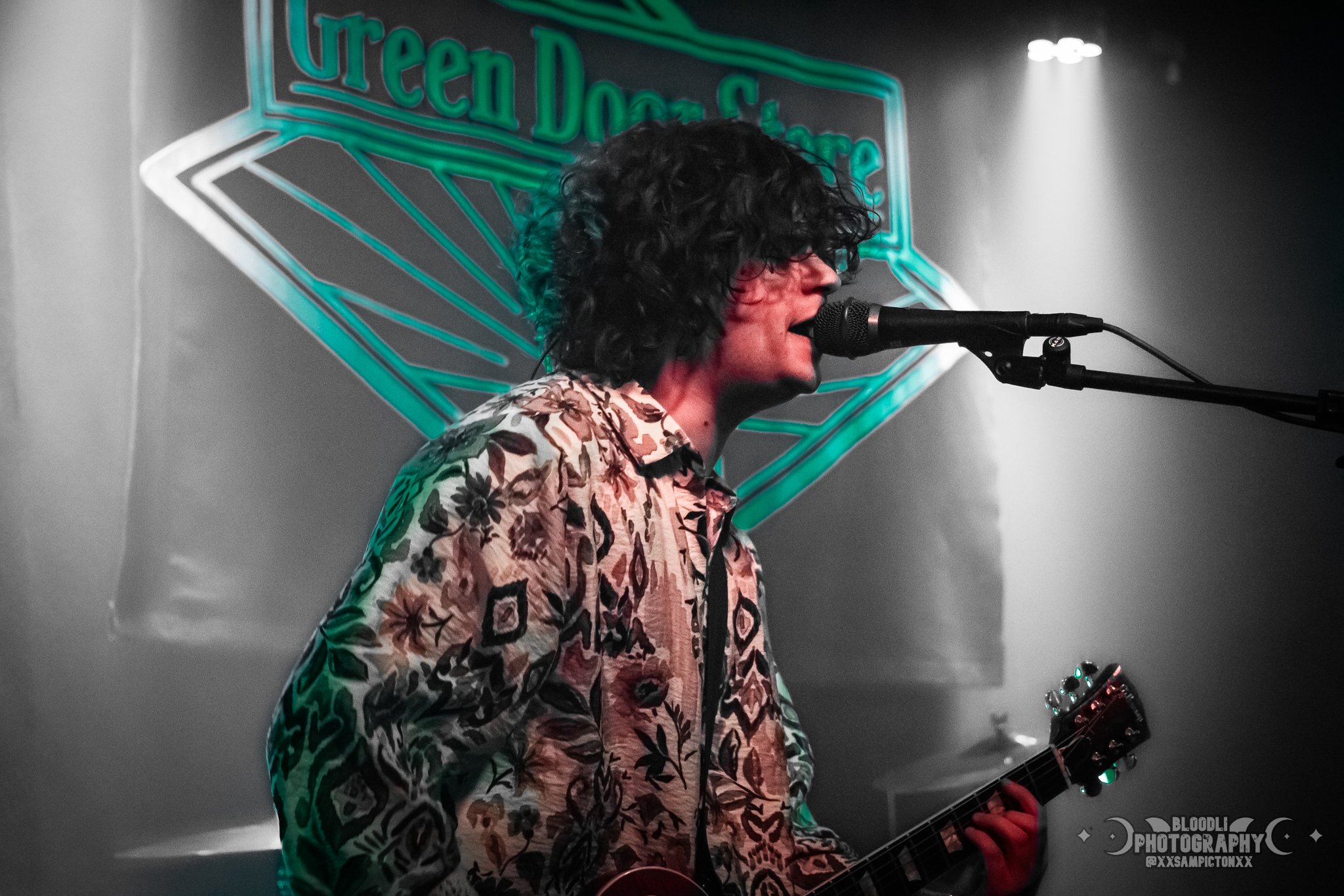 A musician with curly black hair wearing a floral shirt singing into a microphone while playing guitar at a venue. A neon sign in the background reads 'Green Door Store'.