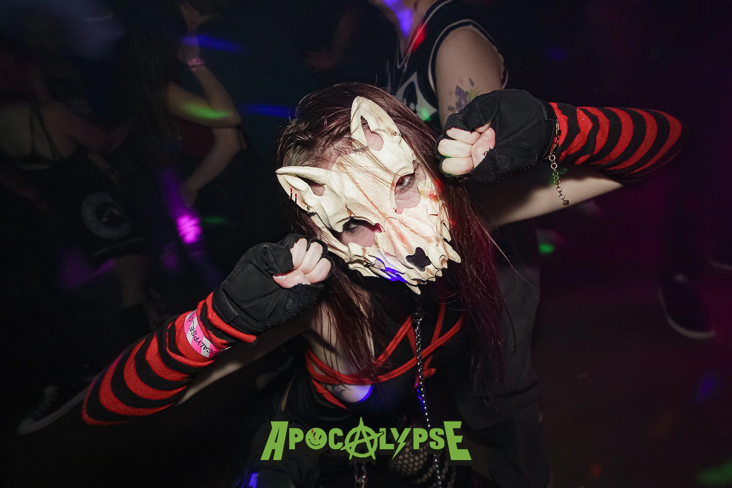 Person wearing a white animal skull mask, black and red striped arm warmers, and black gloves, raising fists in a dance pose at a dark, colorful party with the logo 'Apocalypse' on the bottom.