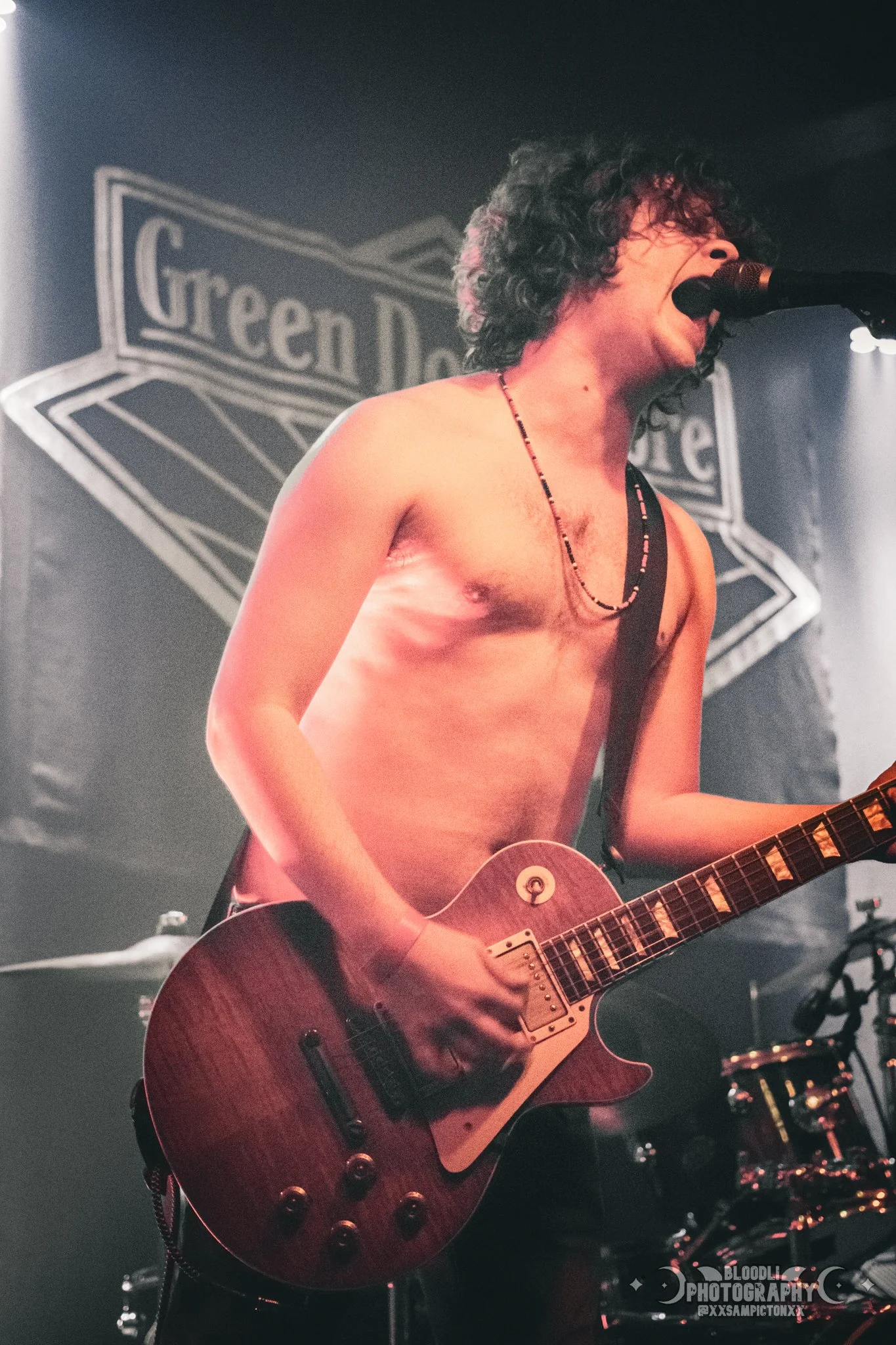 A shirtless male musician with curly hair singing into a microphone while playing an electric guitar on stage, with a 'Green Door' sign in the background.