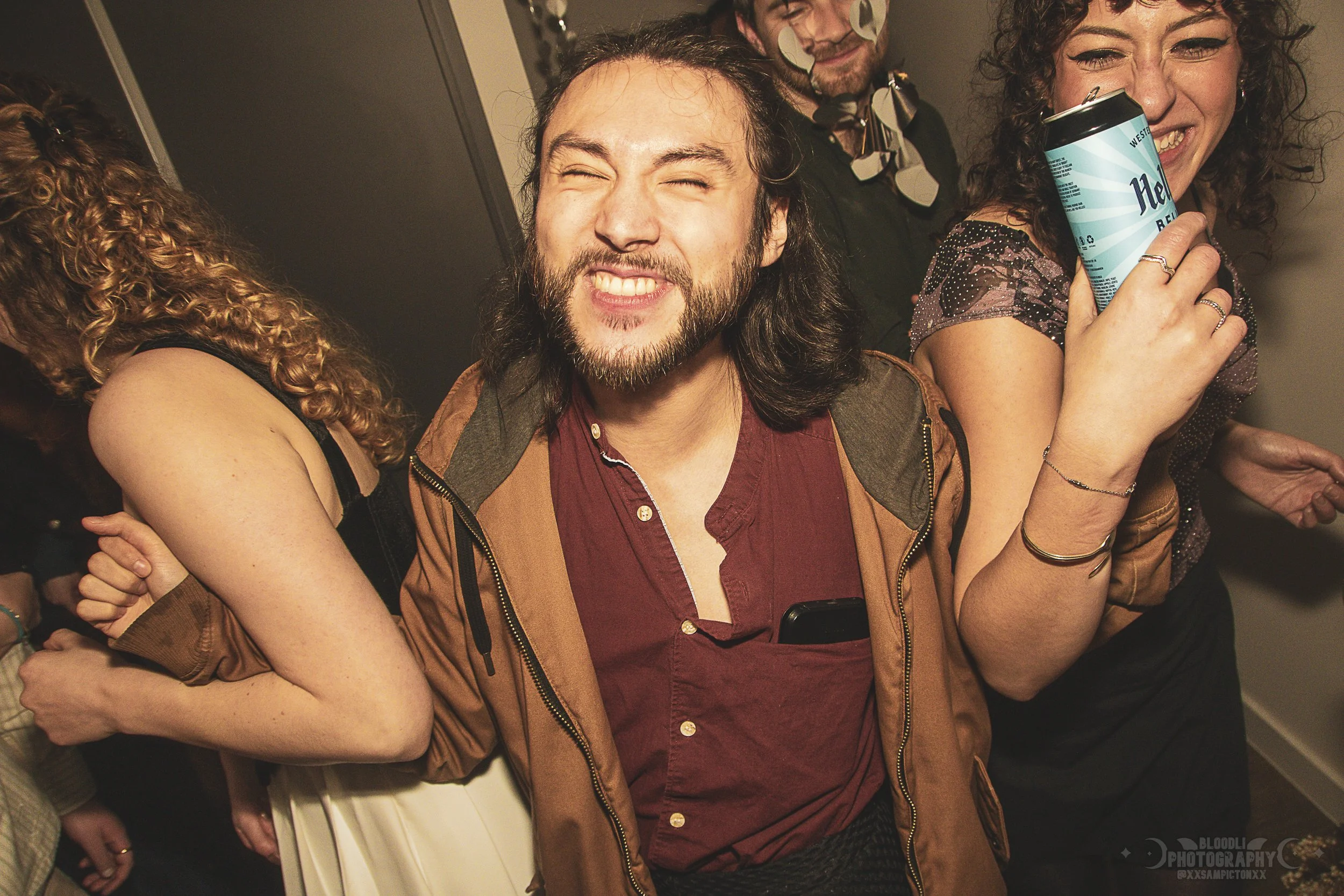 Group of people at a party, smiling and having fun, with one man in the center making a cheerful face and a woman holding a can of beer near her face.