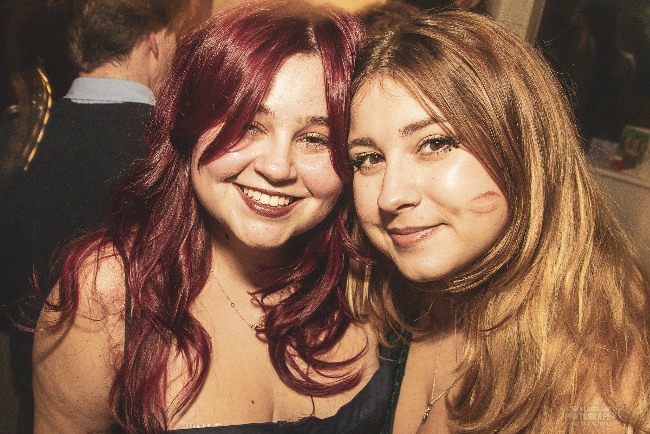 Two young women smiling at a party, one with long red hair and the other with long light brown hair, both wearing necklaces and dressed in dark tops.