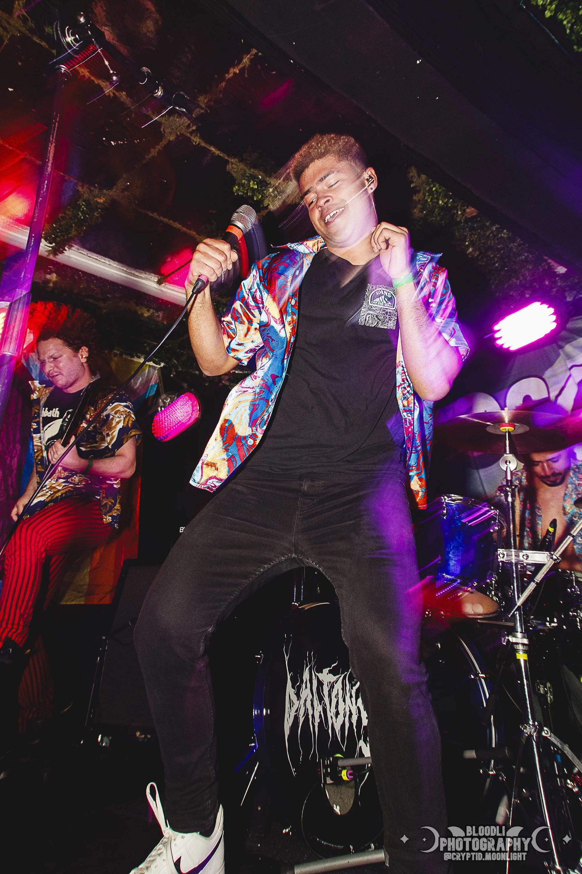 A young man singing into a microphone on stage, wearing a colorful shirt, black t-shirt, black jeans, and white sneakers, with a smile and closed eyes, during a live music performance under colorful stage lights.