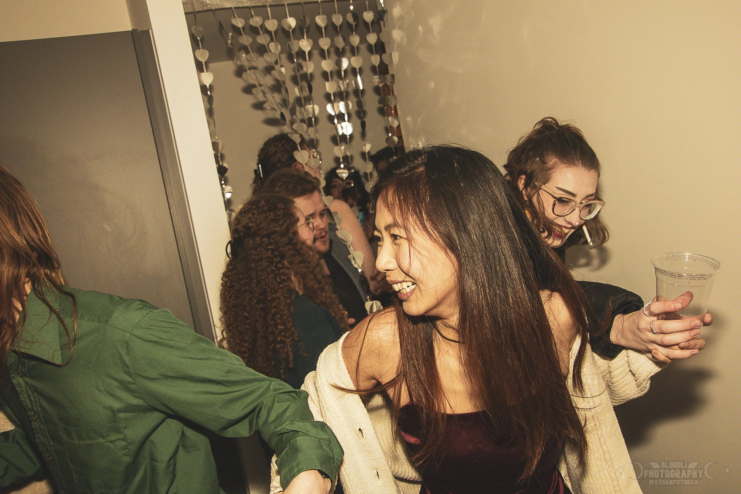 Group of friends at a party, laughing and chatting indoors, with a decorative heart-shaped hanging decoration in the background.