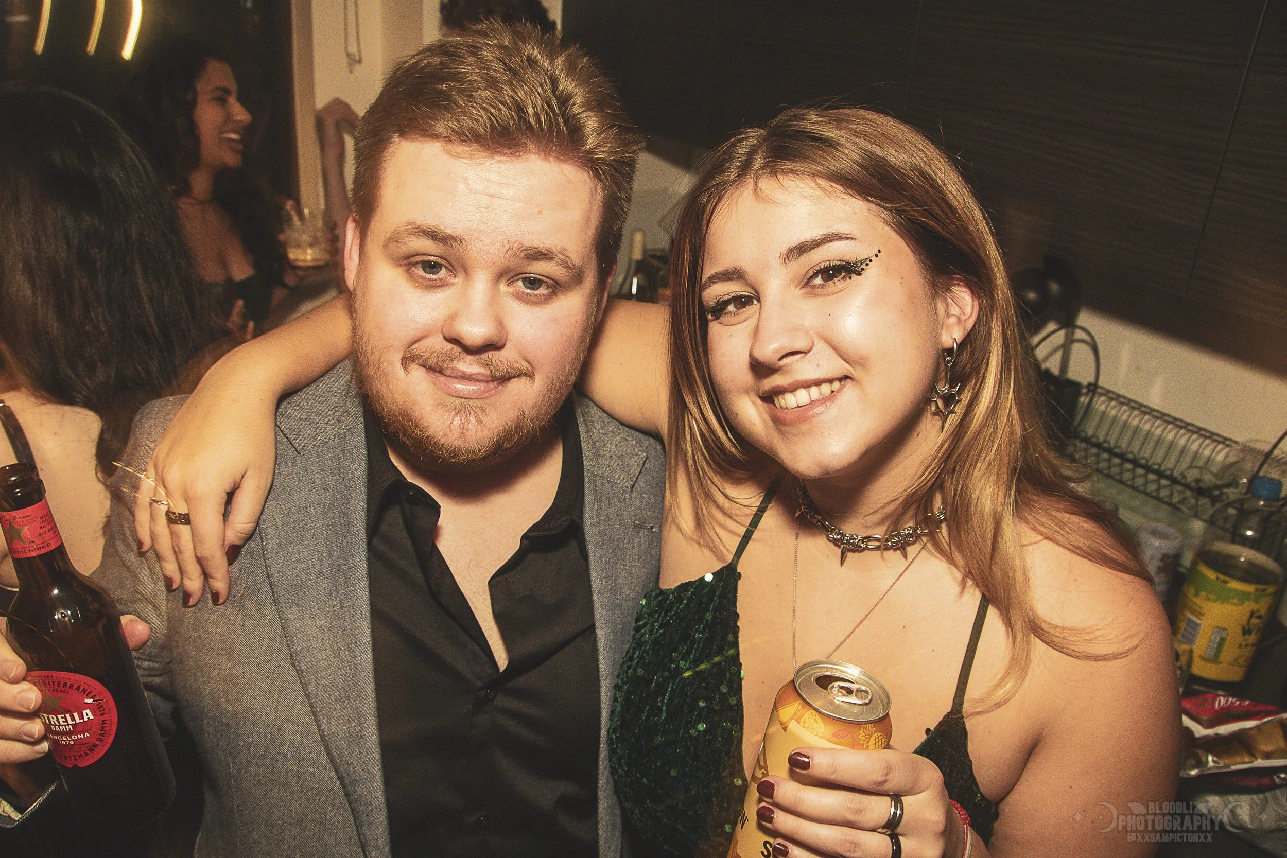 Two people smiling at a party, one man holding a beer bottle and one woman holding a drink can, with other partygoers in the background.