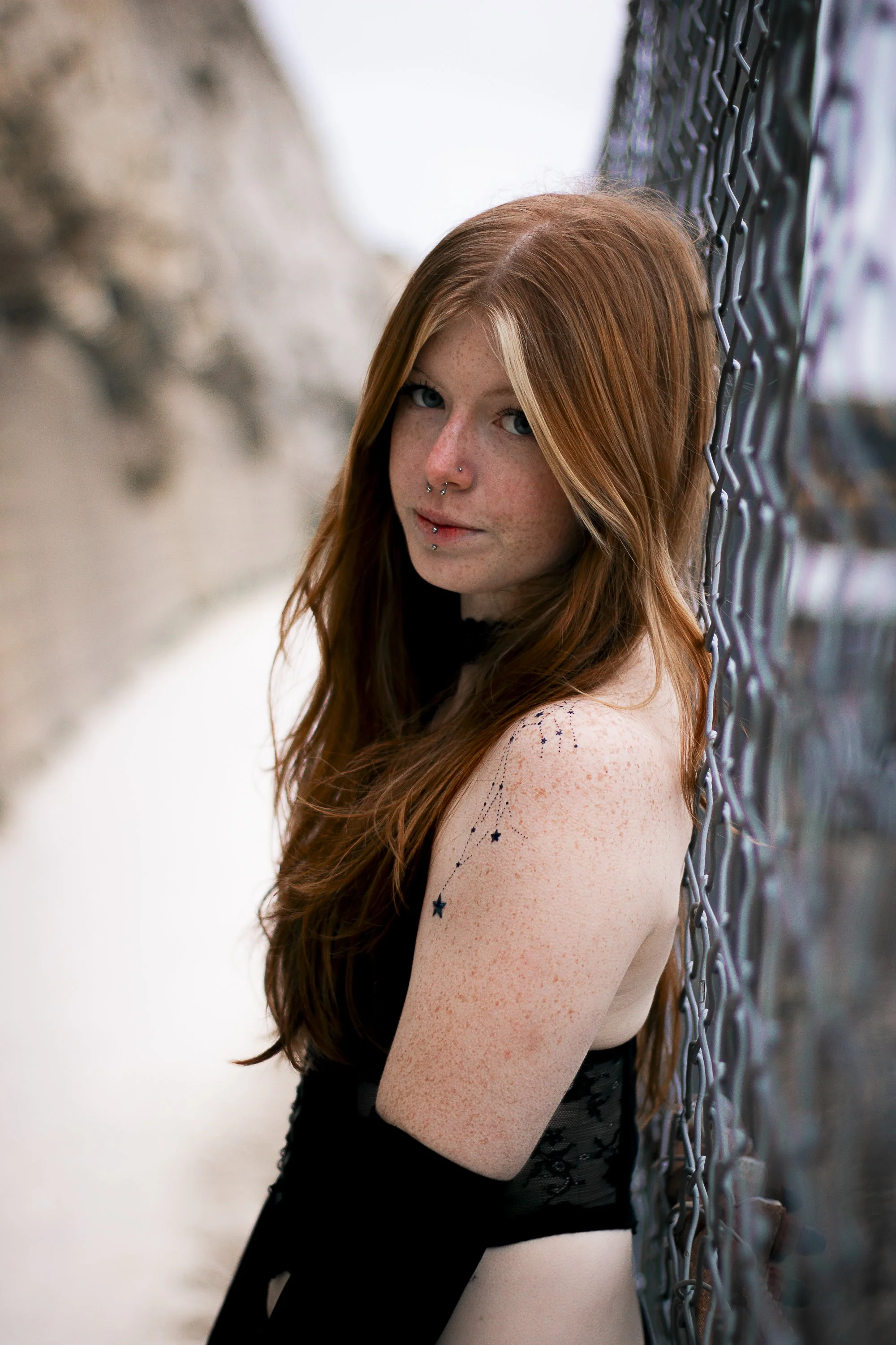 A young woman with long red hair, freckles, and a septum piercing, standing outdoors next to a chain-link fence with snow on the ground and a stone wall in the background.