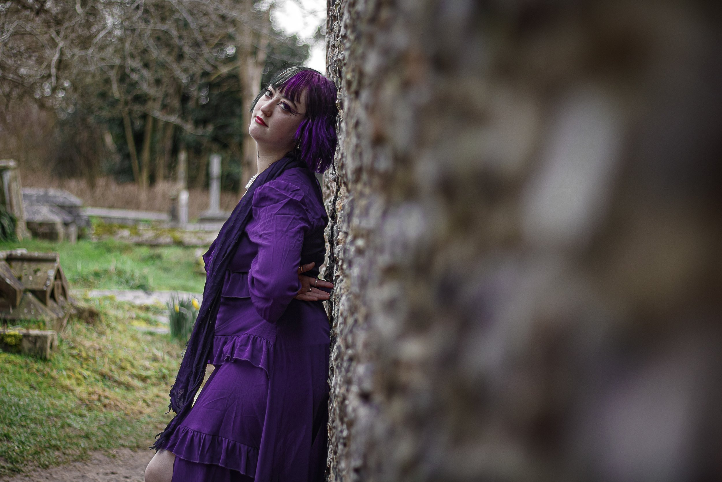 A woman with purple hair and a purple dress leaning against a stone wall outdoors in a garden or park.